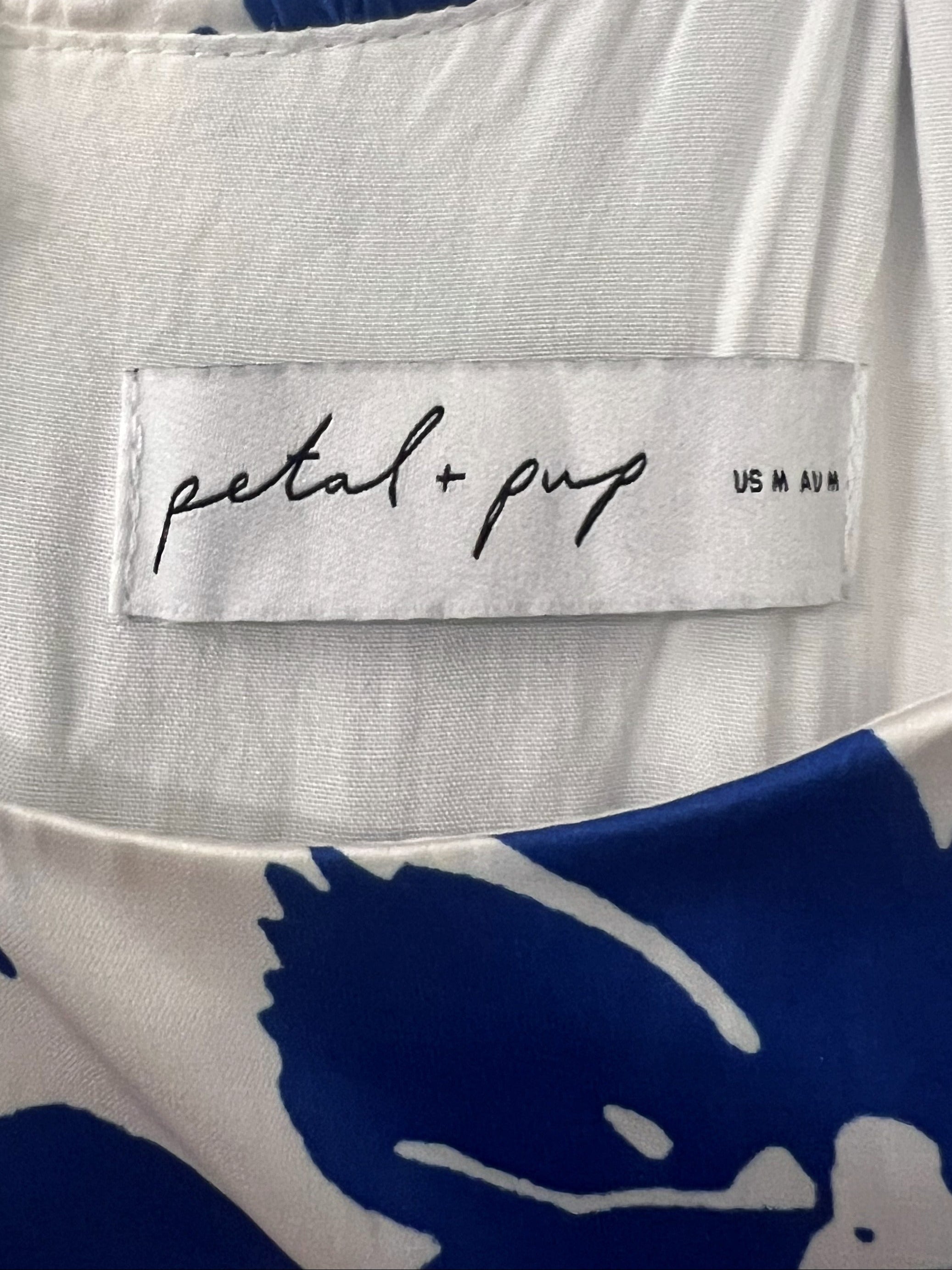 Close-up of a white garment with a blue floral print, featuring a white zipper and a central label that reads 'petal + pup'.