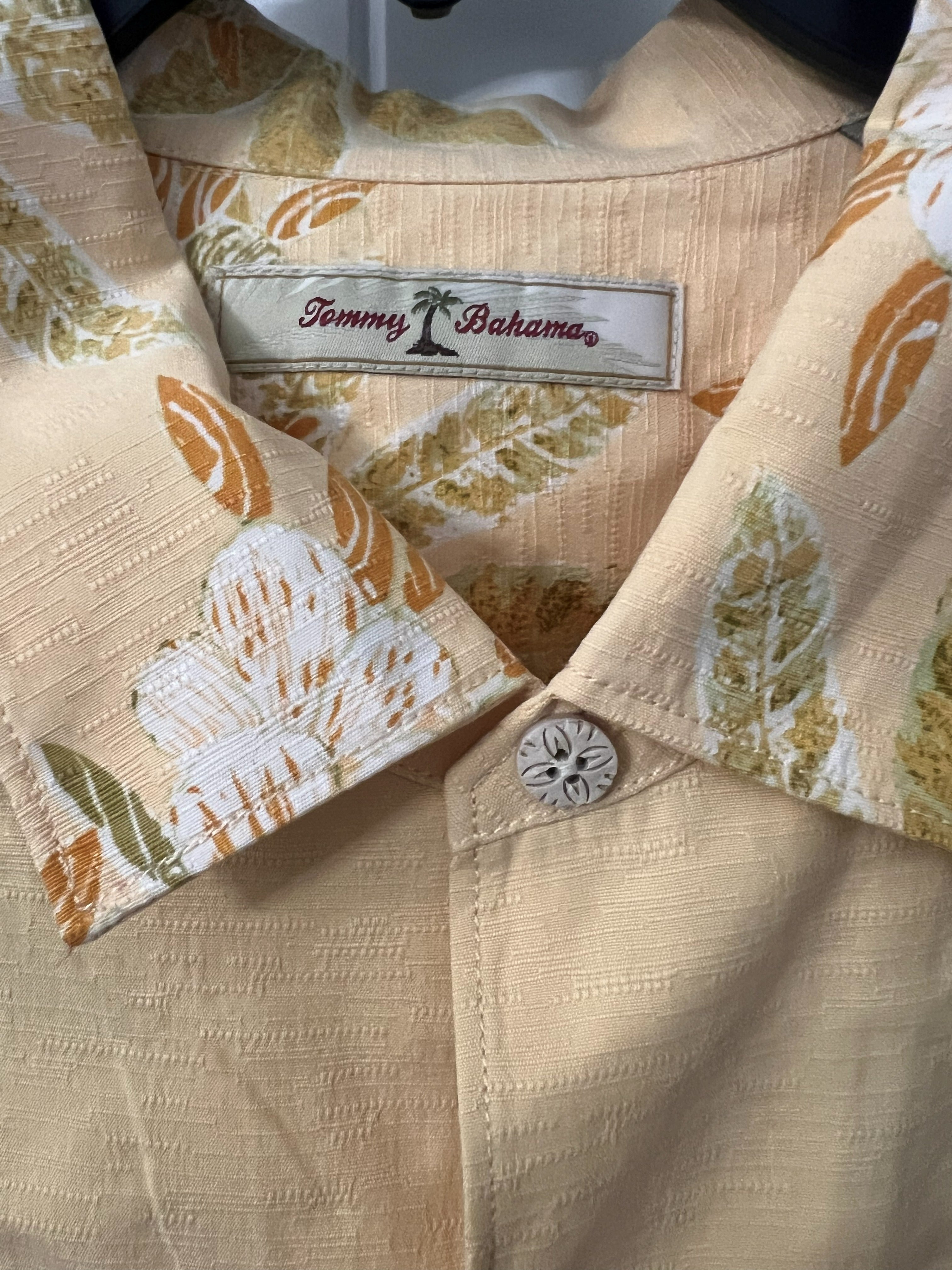 Close-up of a Tommy Bahama short-sleeve button-up shirt with a textured cream base and a tropical floral leaf print in orange, olive green, and white. The Tommy Bahama brand label is visible on the collar, and a decorative shell-style button is featured on the placket.