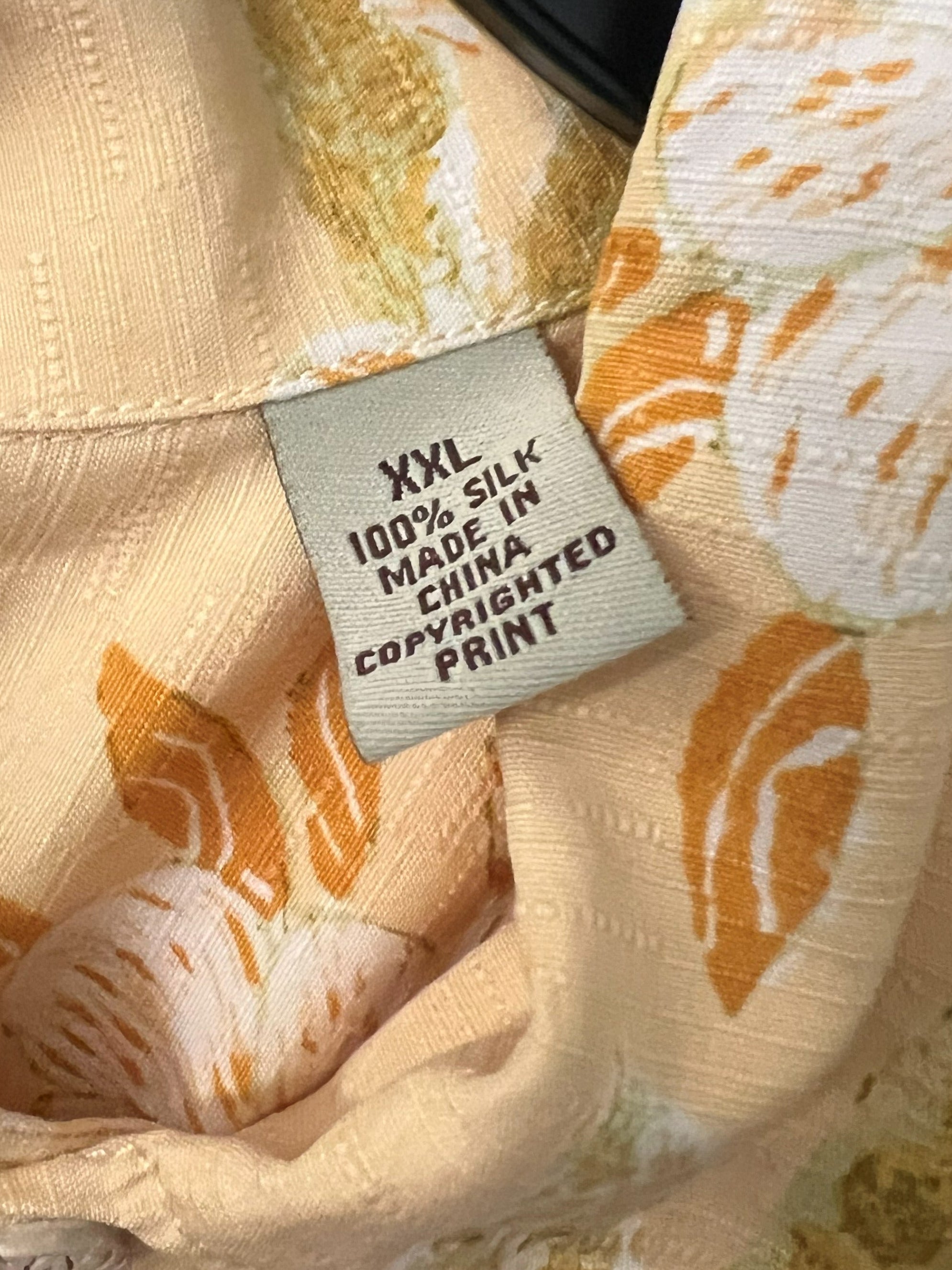 Close-up of a light beige and orange floral printed shirt on a black hanger, showing a fabric care label that reads 'XXL 100% SILK, MARIJE CHINA CHIKARA CONFIATED PRINT'.