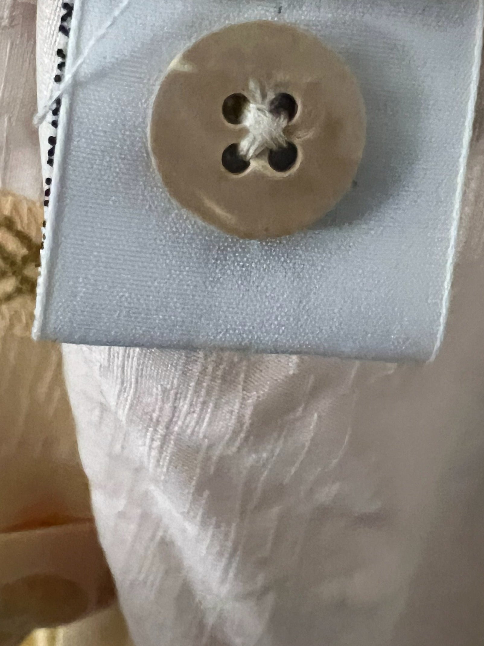 Close-up of a light beige fabric garment with a large round beige button attached to a white fabric tag, showing textured woven material and a hint of gold patterned trim.
