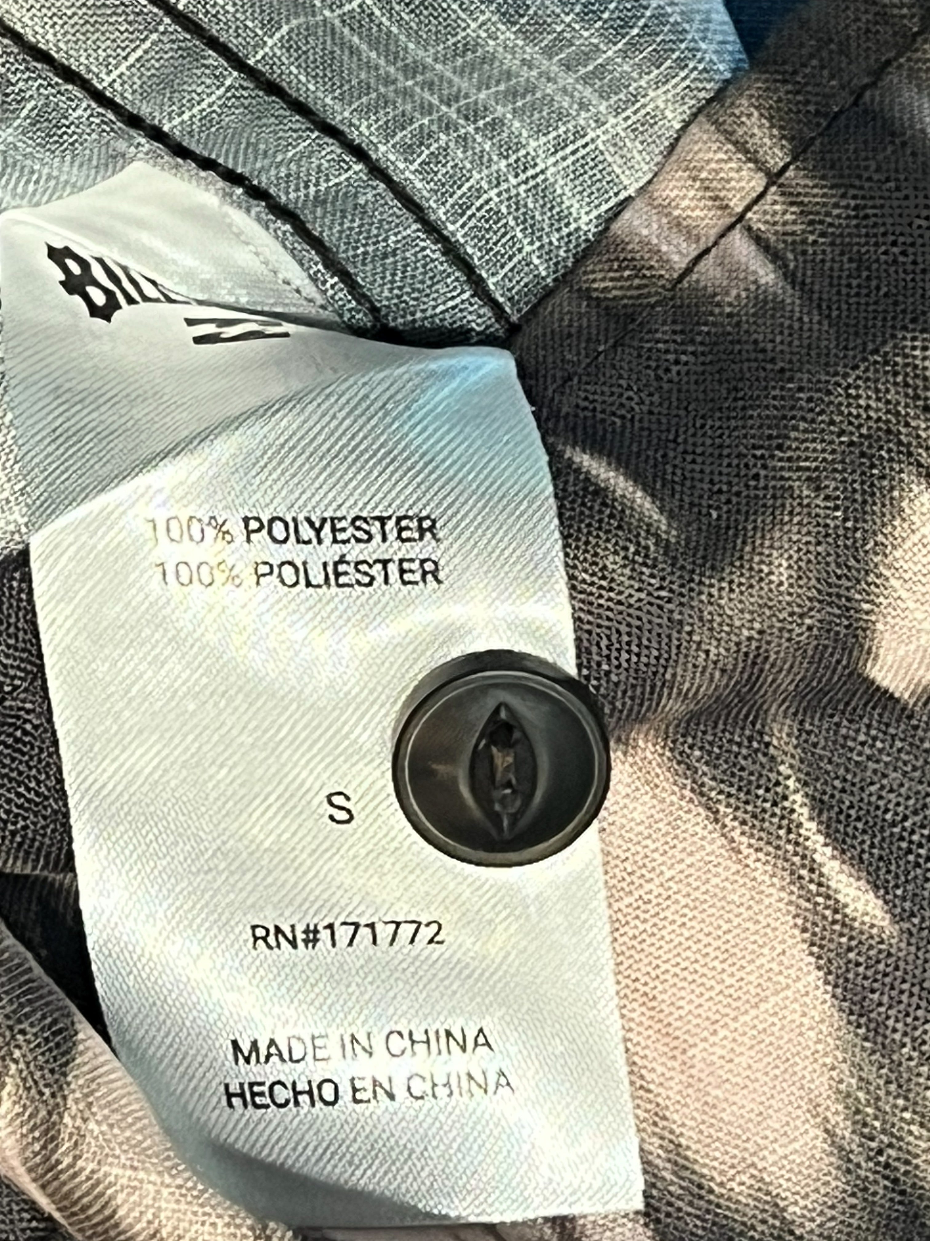 Close-up of a garment's inner neck label made of light blue-gray polyester, sewn onto a gray textured fabric with black piping. The label shows size S, '100% POLYESTER', 'MADE IN CHINA', and a black circular button closure.