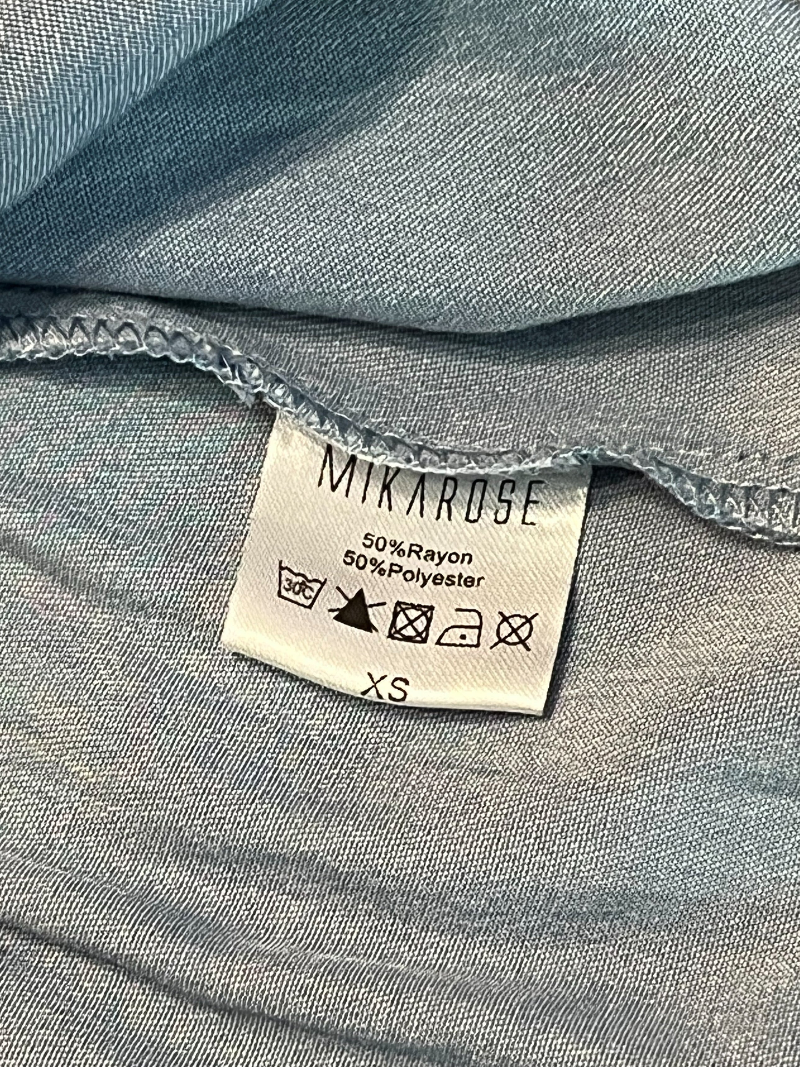 Close-up of a light blue-gray fabric garment showing a sewn-on care label that reads MIKAROSE.