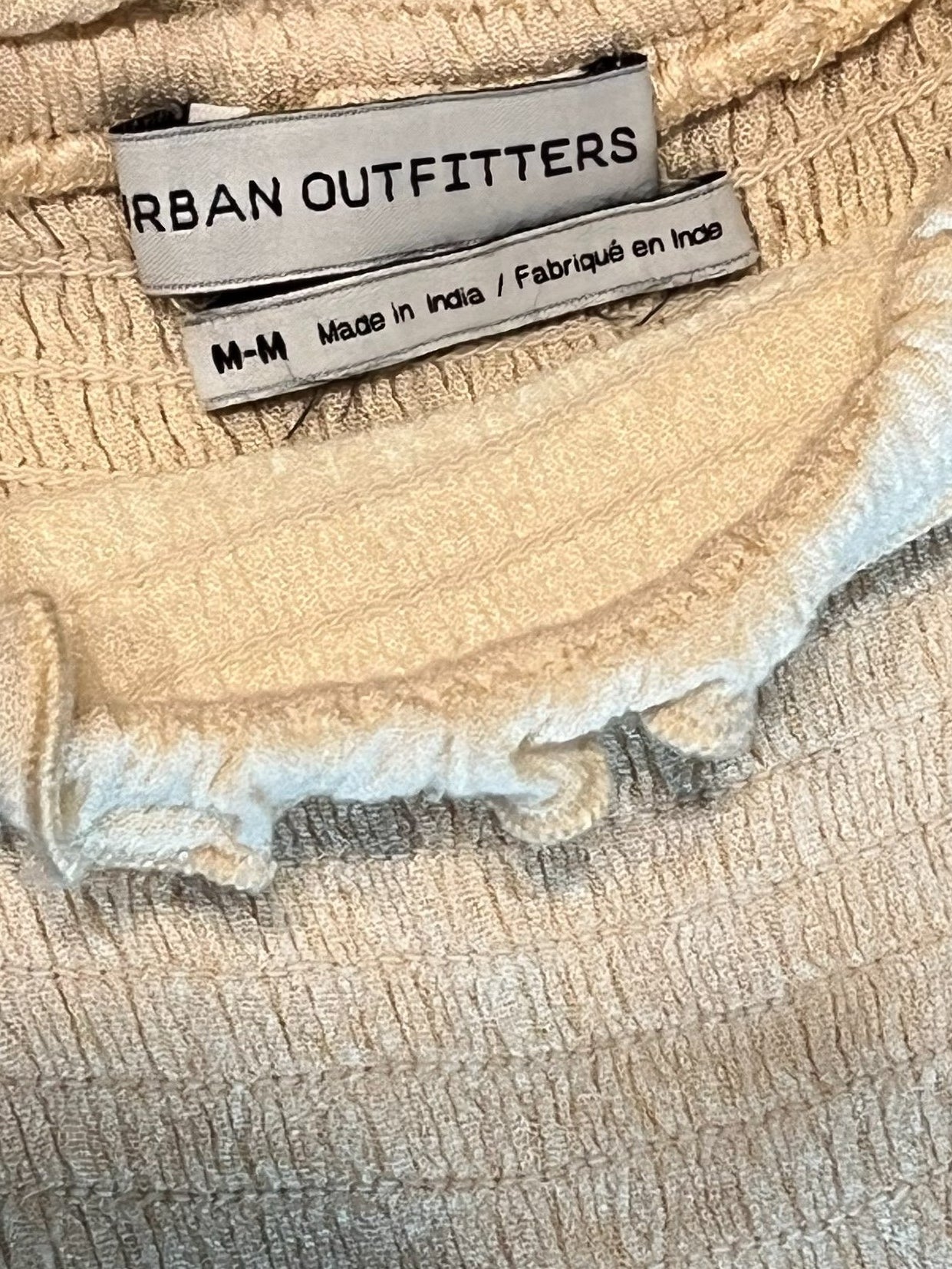 Close-up of a cream-colored textured garment with ruffled trim, showing a sewn-in label that reads Urban Outfitters.