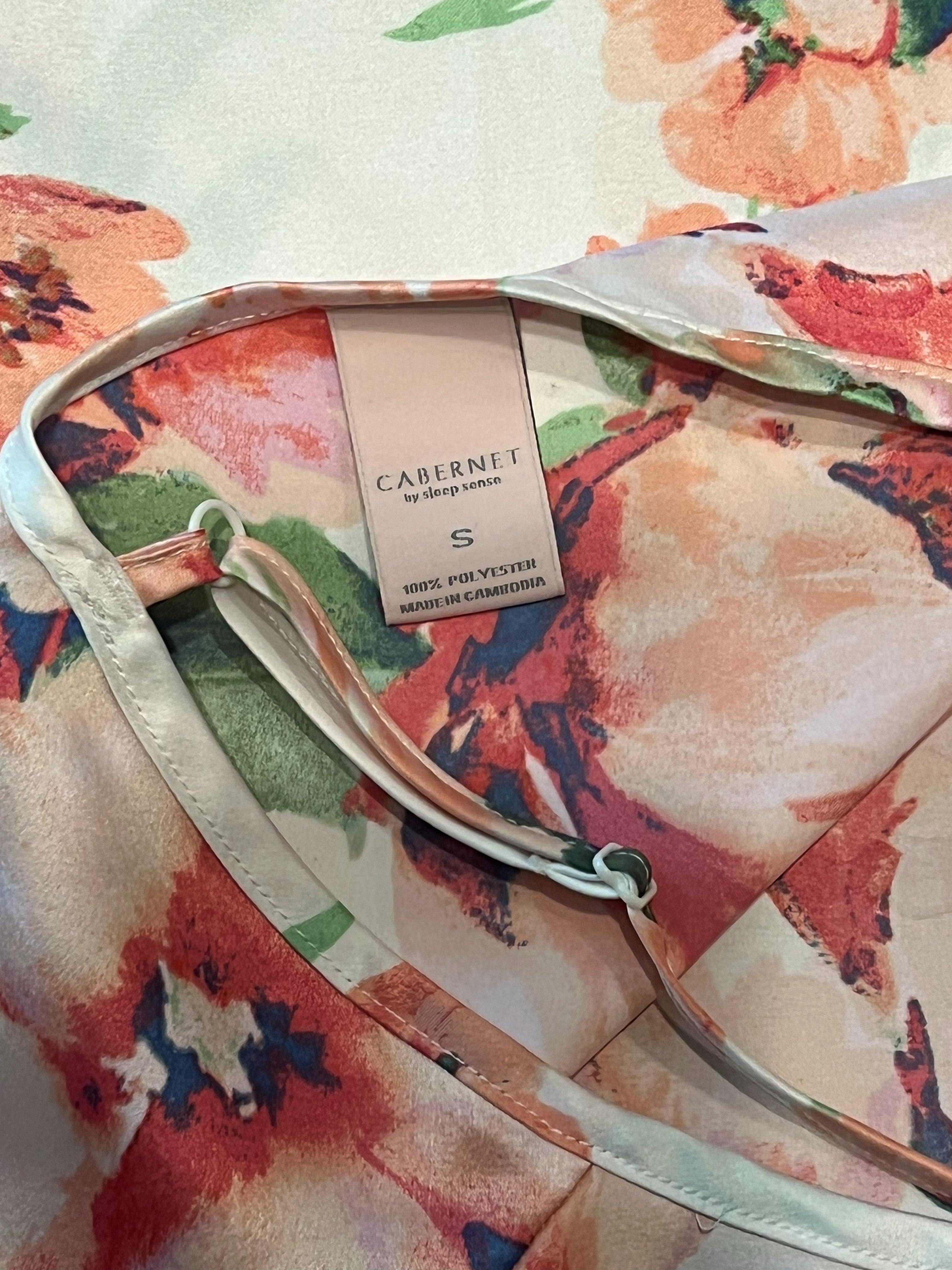 Close-up of a floral-patterned camisole top with a visible neck label reading CABINET by Sarah Wells. The fabric has a soft blush, peach, orange, and green floral print with a pale pink satin-like trim.