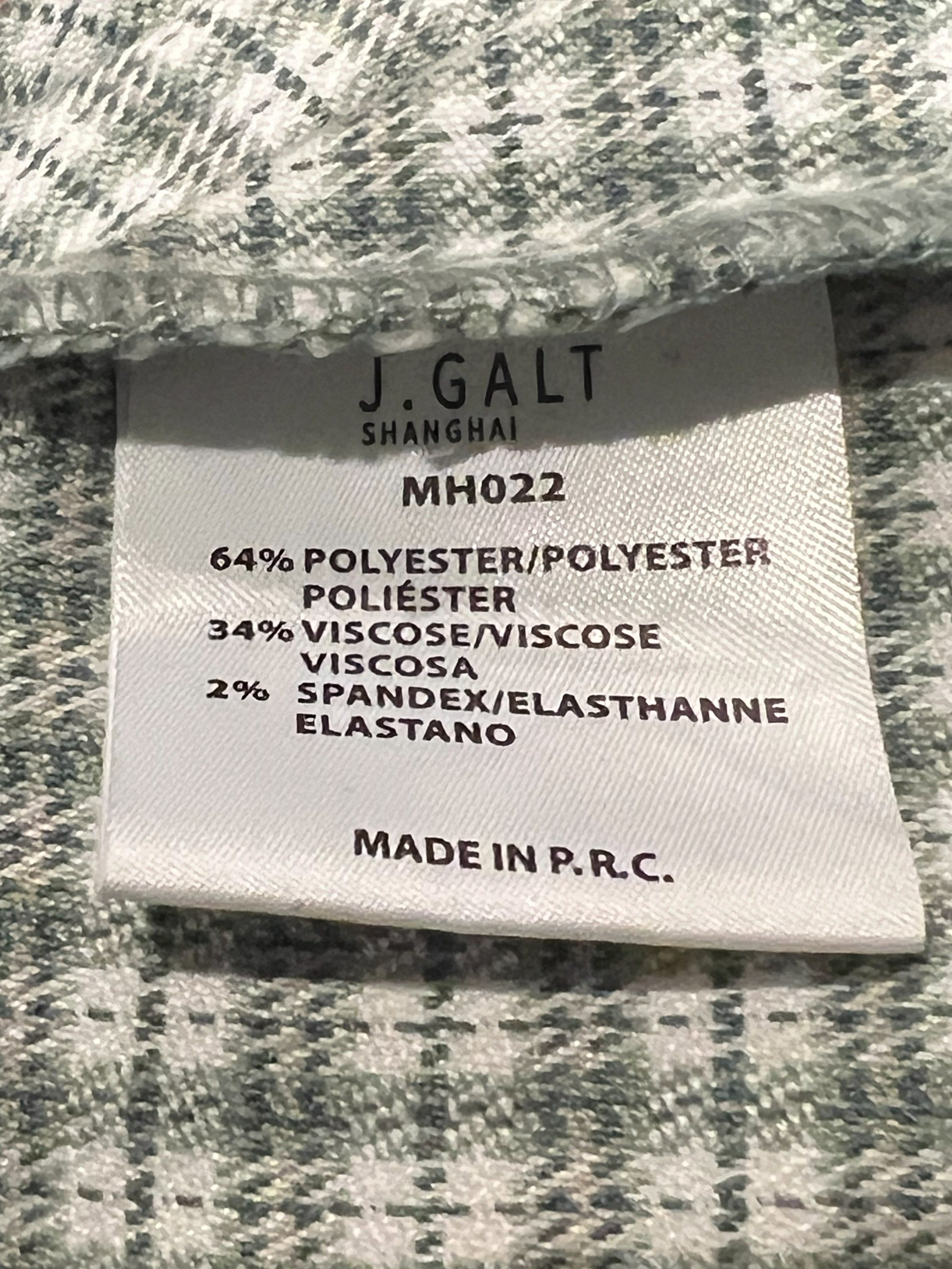 Close-up of a woven fabric label sewn onto a green and white plaid textile, showing text including 'JGALT' and material composition details.