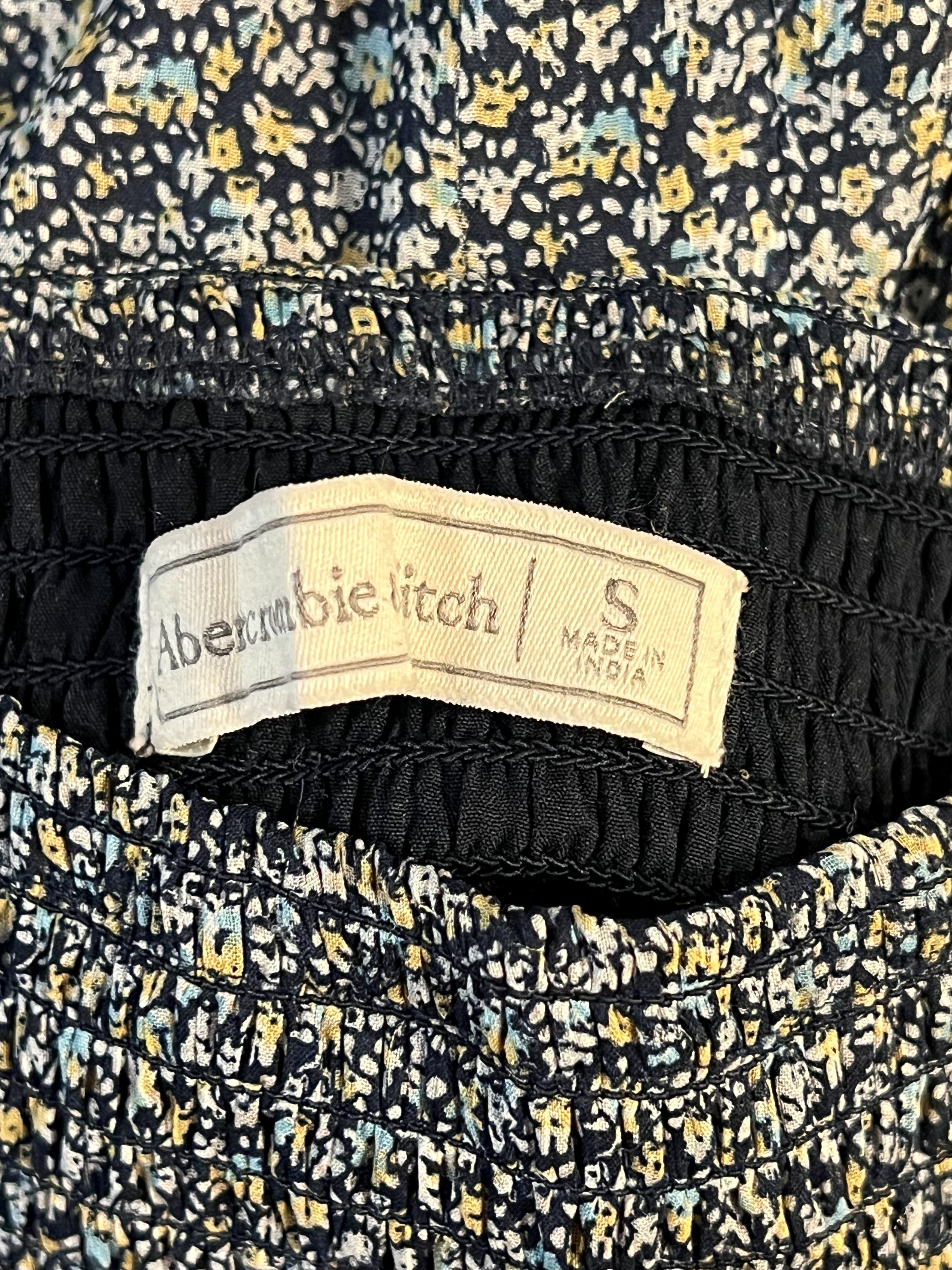 Close-up of a garment's inner waistband showing a cream-colored fabric brand label, with a black elastic band and a black, yellow, and blue floral patterned fabric visible.