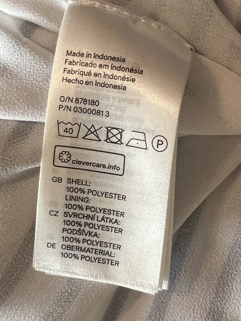Close-up of a garment's interior label sewn onto a light beige or off-white fabric, held by a hand. The label contains text and care instruction symbols.