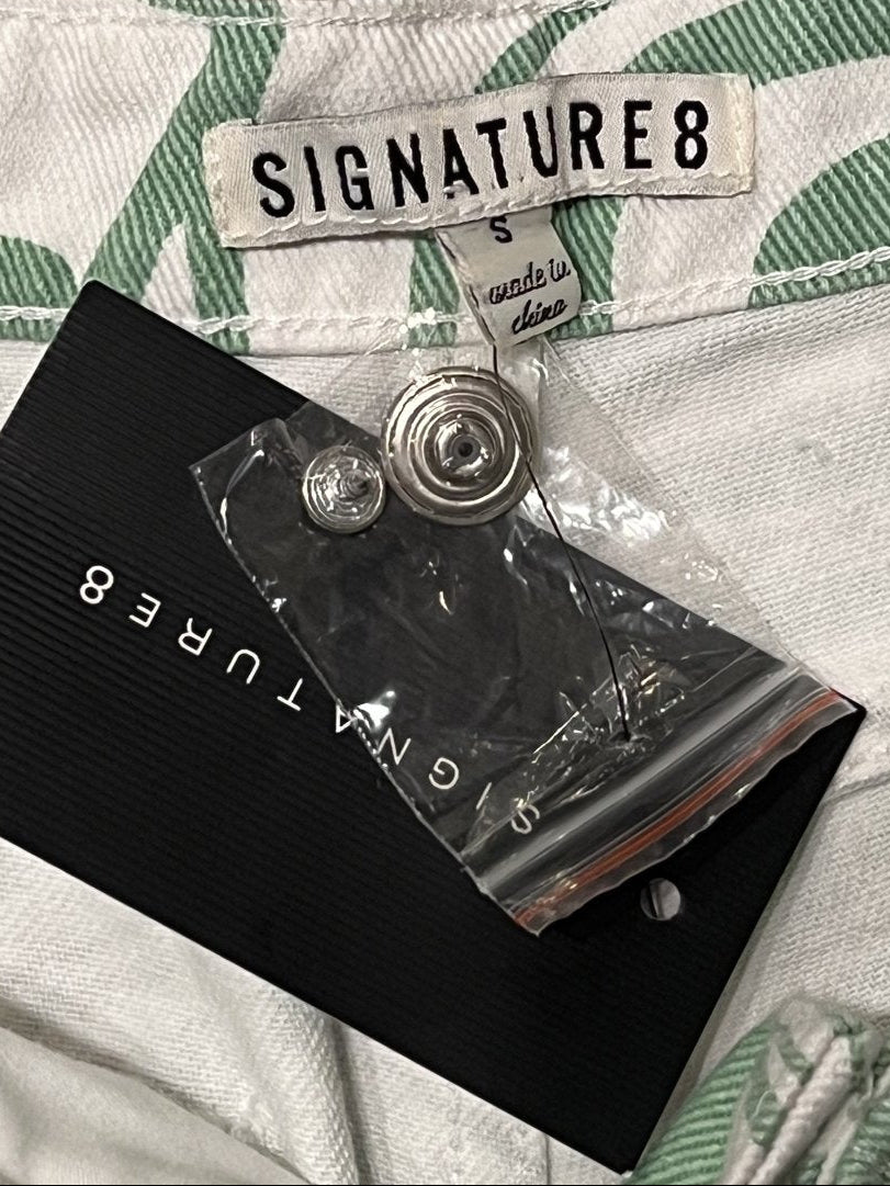 Close-up of a garment tag reading SIGNATURE8 on a white fabric with green leaf pattern, next to a black hangtag and a small clear plastic pouch containing a metal button.