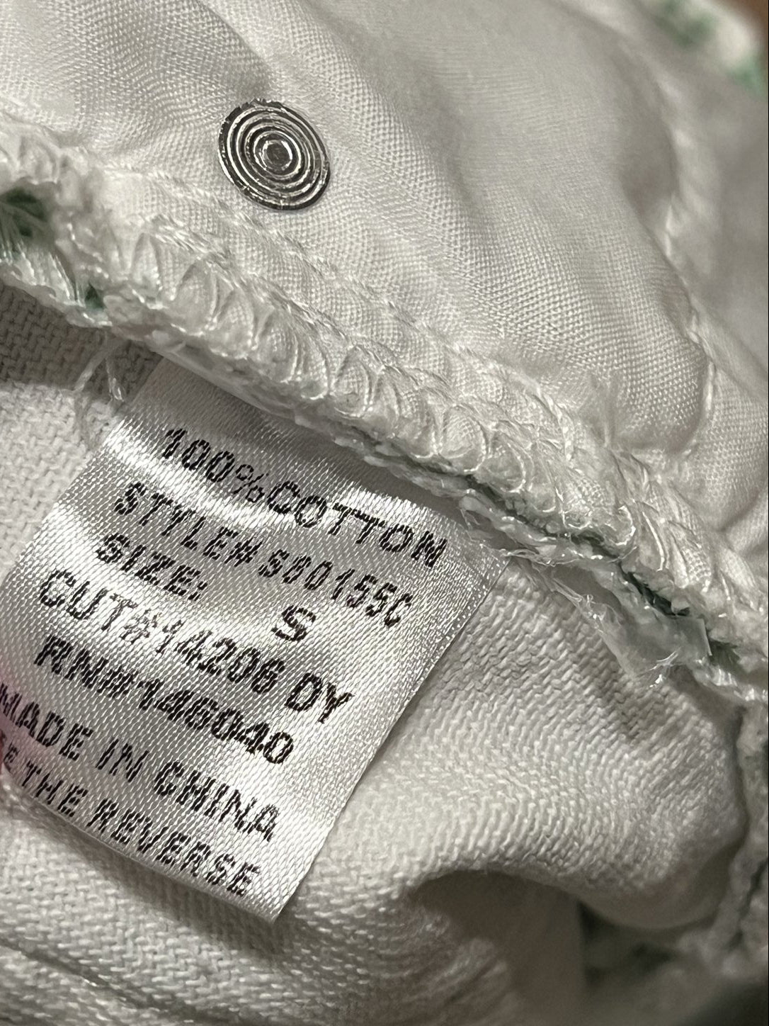 Close-up of a white cotton garment label sewn onto a white fabric item, showing text including '100% COTTON', 'SIZE S', and 'MADE IN CHINA'.