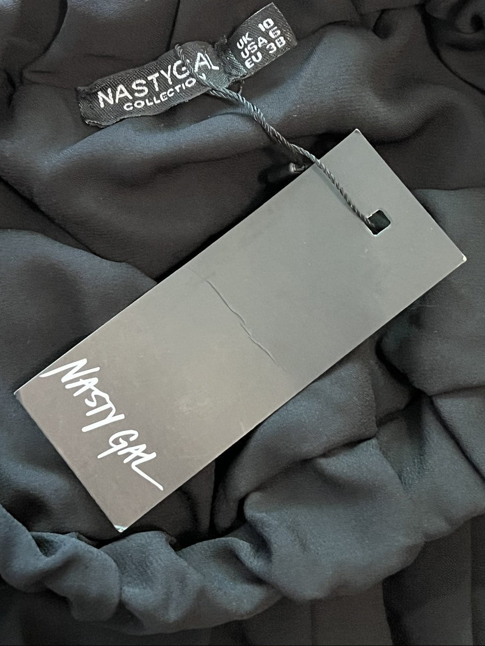 A close-up of a dark gray garment with a large blank gray hangtag and a smaller black size/brand tag reading 'NASTY GAL'.