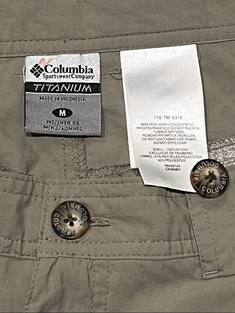 Close-up of the waistband of olive green Columbia pants, showing the white size tag, the black and white WINDRUSH technology label, and the metal button closure.