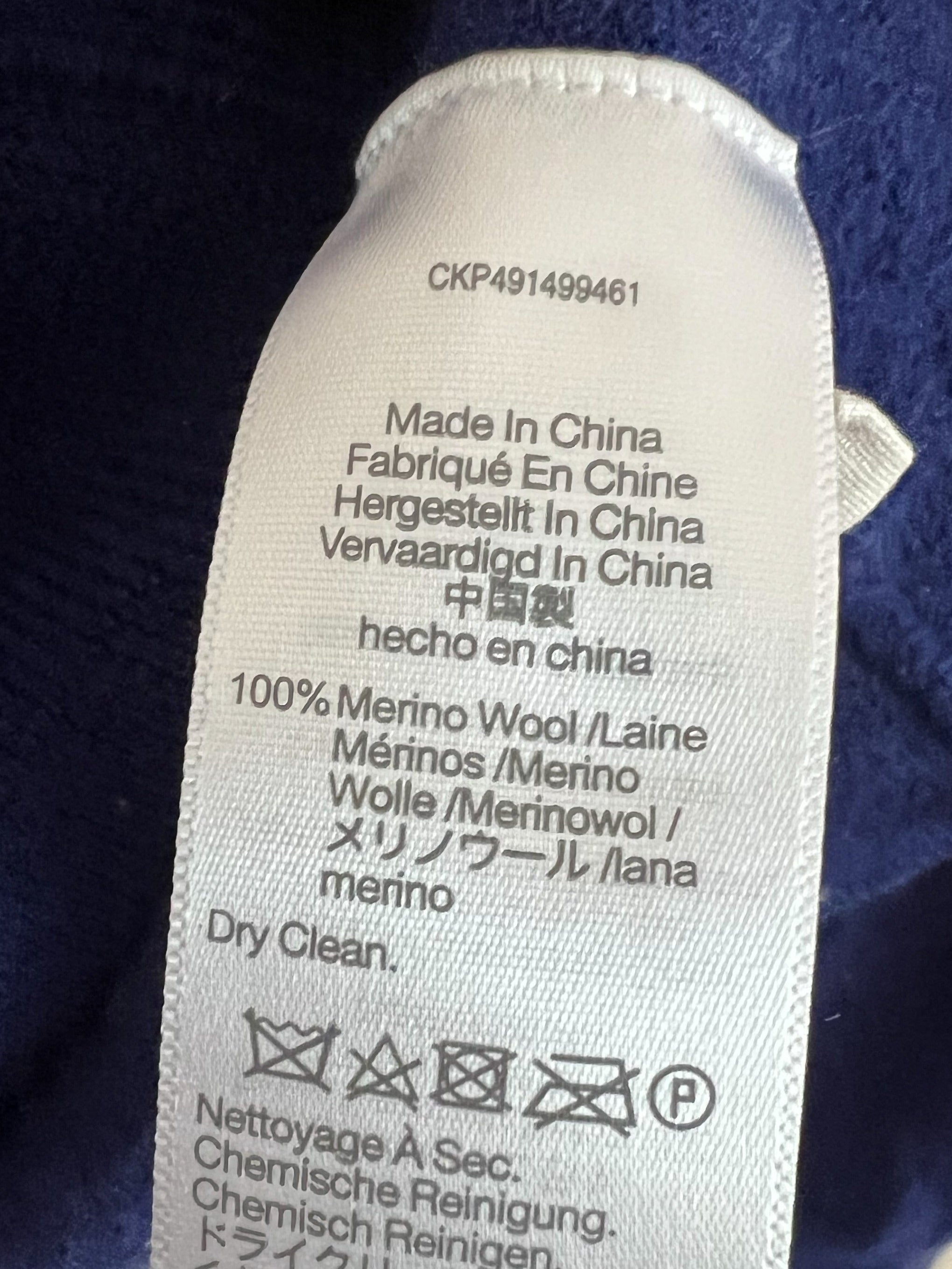 A close-up of a white garment care label sewn onto a dark blue textured fabric, showing text including '100% Merino Wool / Laine' and care symbols.