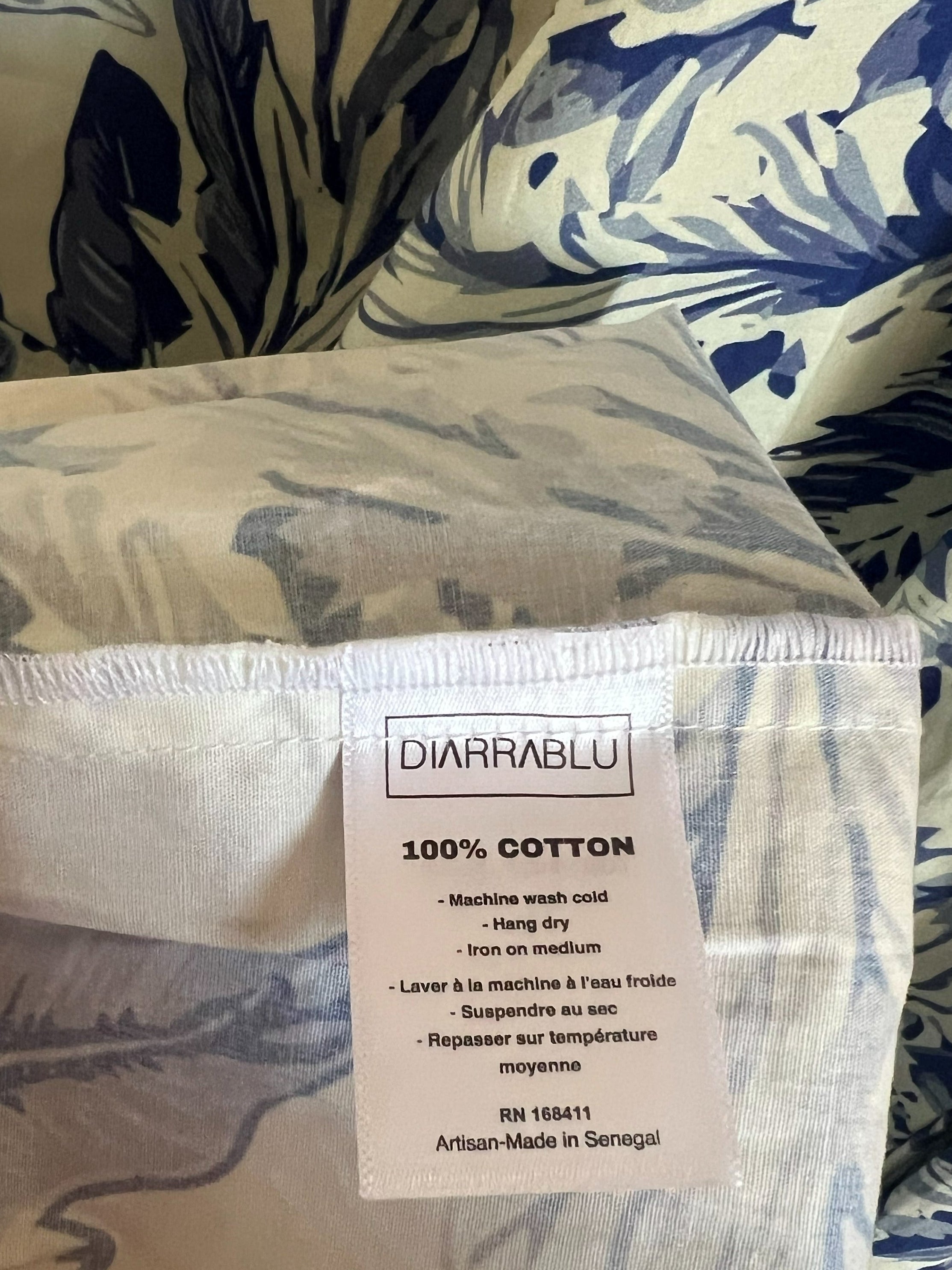A close-up of a folded cotton textile with a blue and beige botanical leaf print, showing a white sewn-in label that reads 'DIARRABLU' and '100% COTTON'.