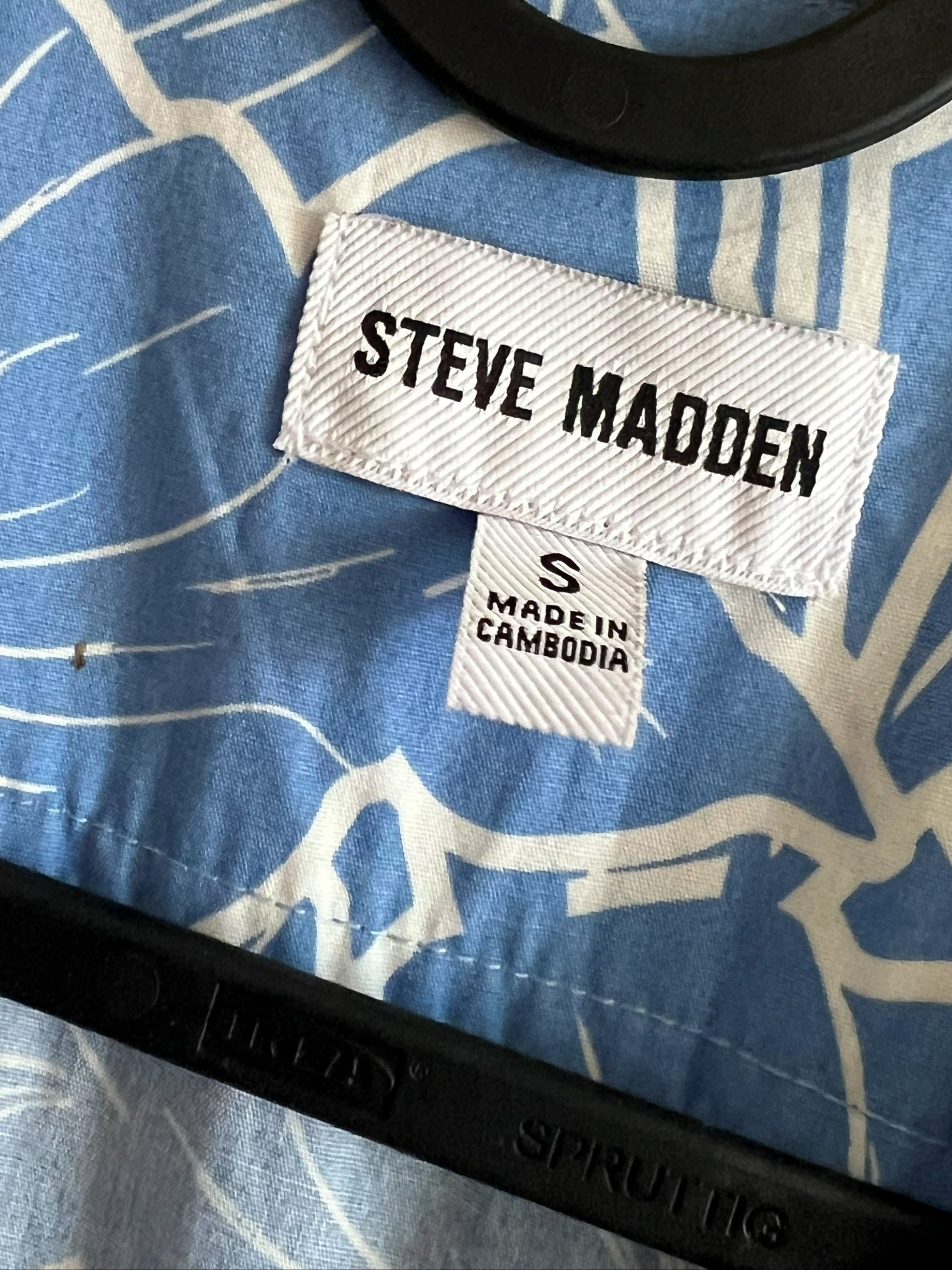 Close-up of a blue and white patterned shirt on a black hanger, showing a visible Steve Madden brand label and a smaller Made in Cambodia tag.