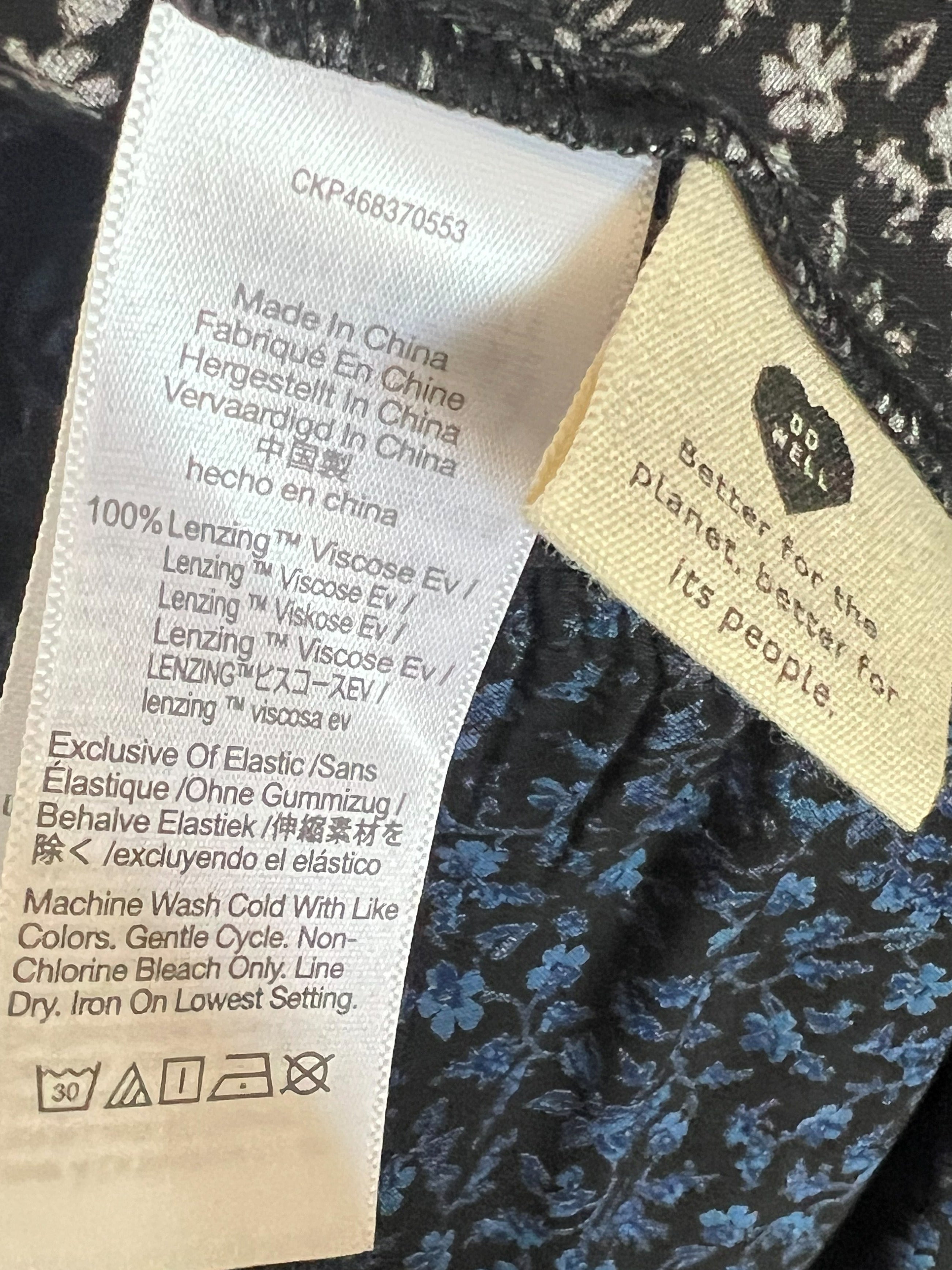 Close-up of a garment's interior label area showing a white care tag with text and a beige brand tag reading 'Better for the planet, style its people.' with a black heart logo.