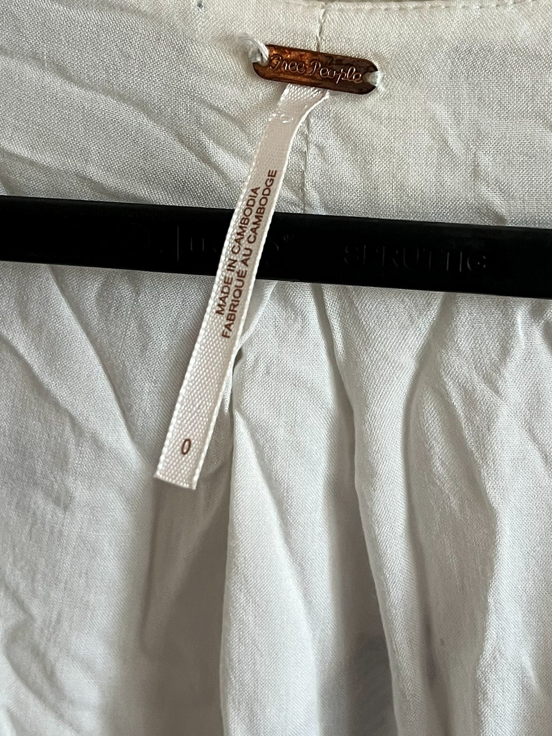 Close-up of a white garment hanging on a black hanger against a textured beige wall, with a visible brand tag and size tag attached at the neckline.