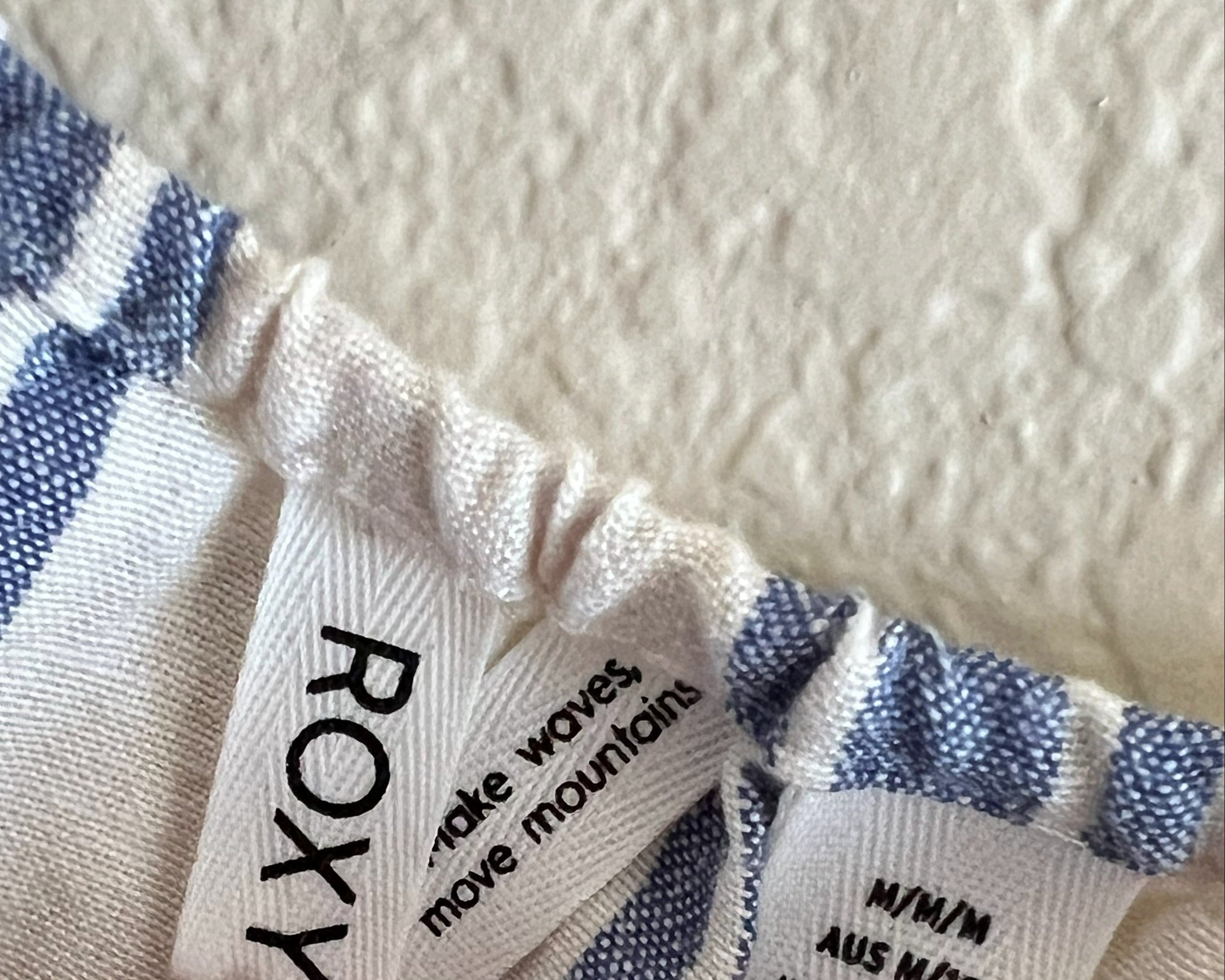 Close-up of a striped fabric garment with visible sewn-in labels reading ROXY and size M/M, with a person's fingers pointing at the labels.