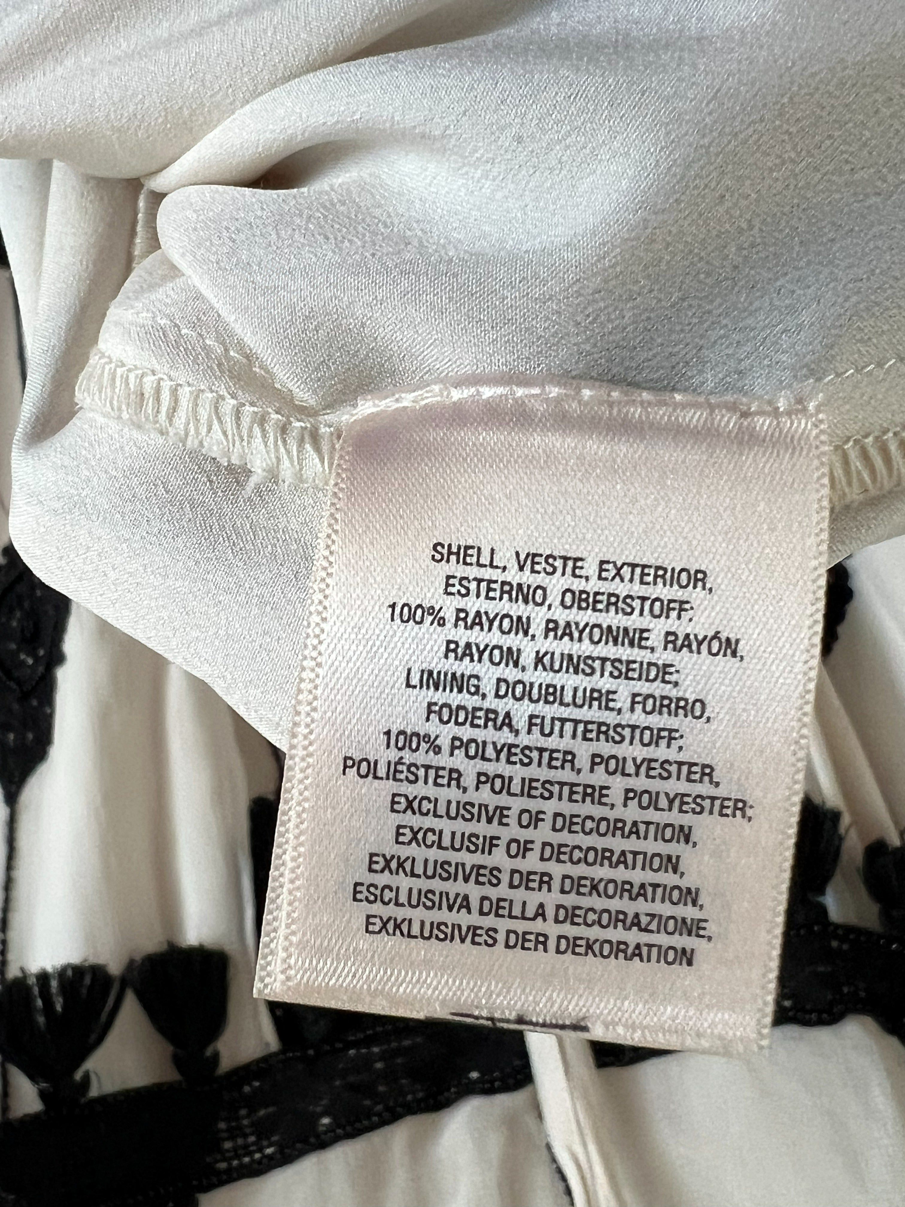 Close-up of a sewn-in fabric care label on a white or off-white garment, showing text about materials and care instructions. Black tassel trim is visible at the top.