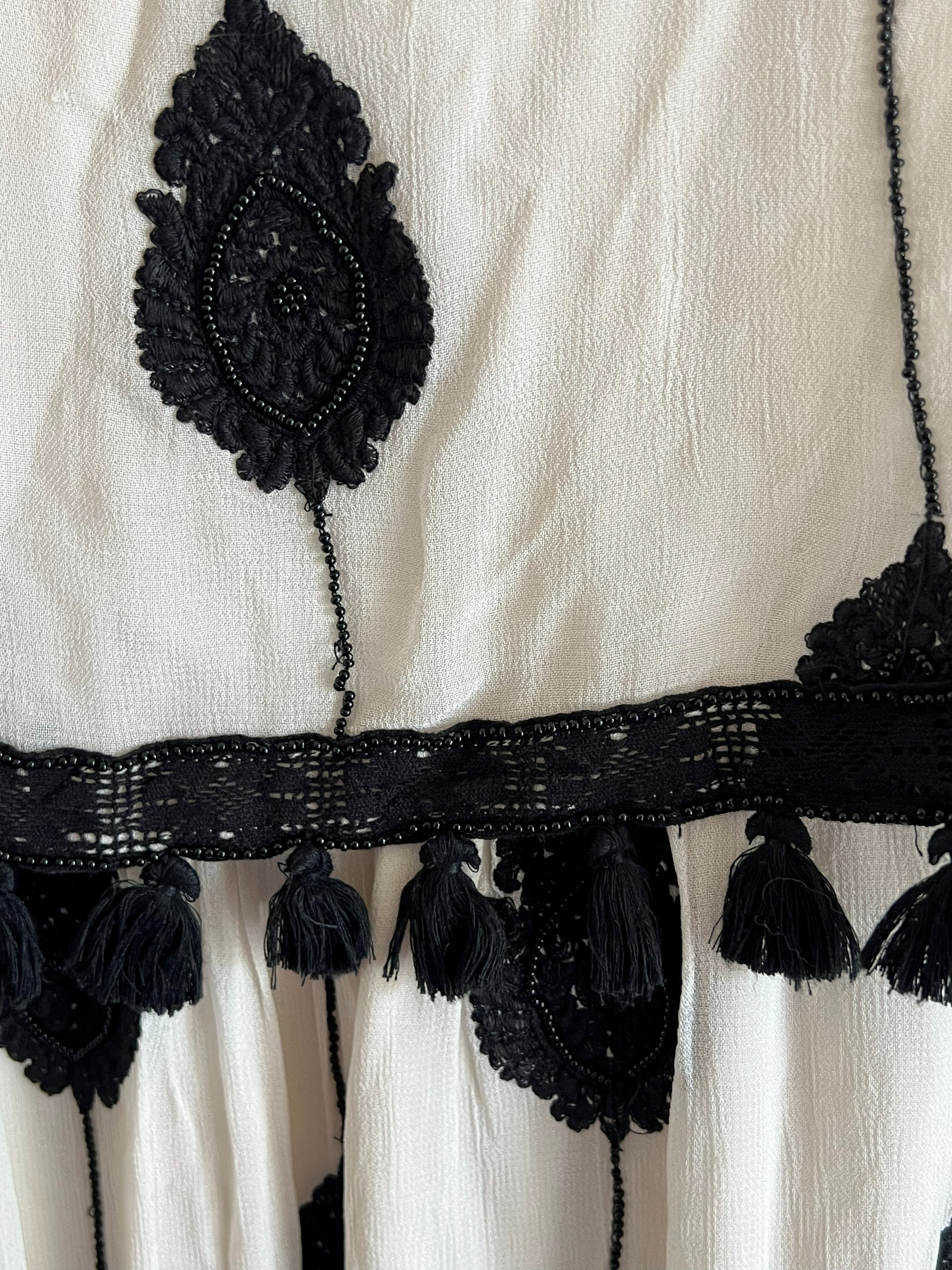 Close-up of a white textured fabric garment featuring black lace applique, a decorative black lace band, and black tassels.