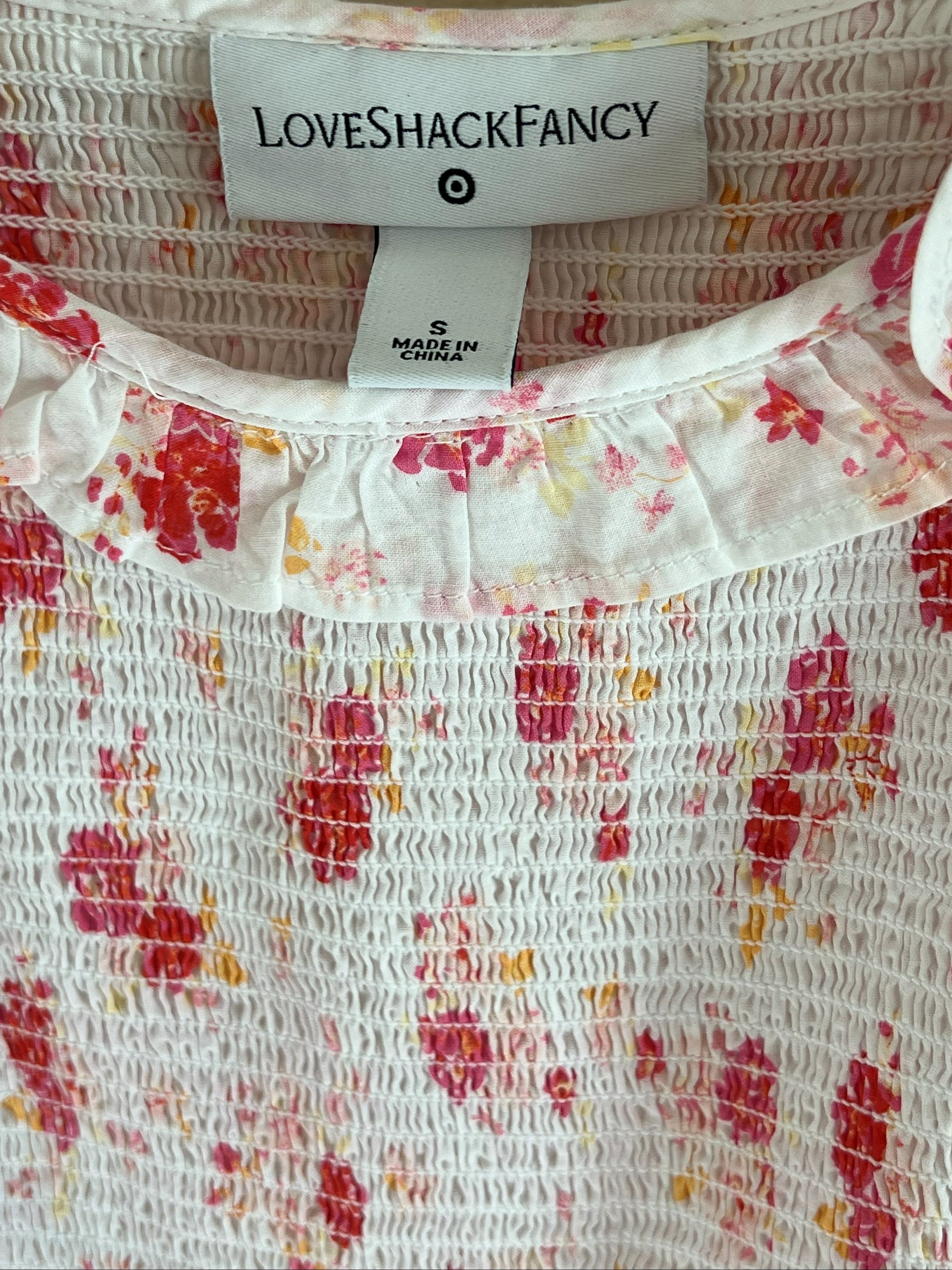 Close-up of a white smocked blouse with a red and orange floral pattern, featuring a ruffled collar and a visible 'LOVESHACKFANCY' brand label.