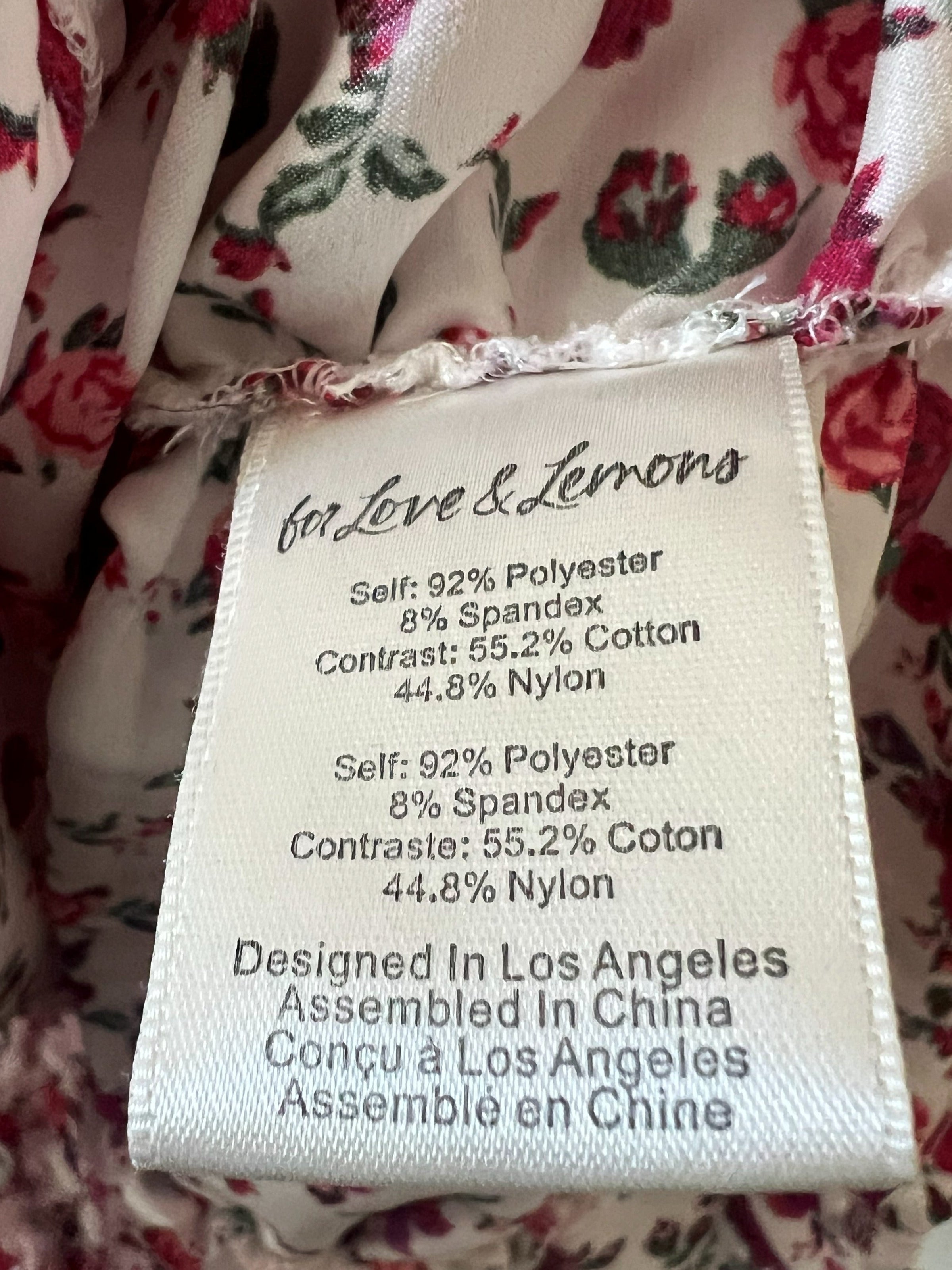 Close-up of a floral garment's interior label reading 'in Love & Lemons' with fabric composition details and manufacturing text, surrounded by pink and green floral print on a light base.