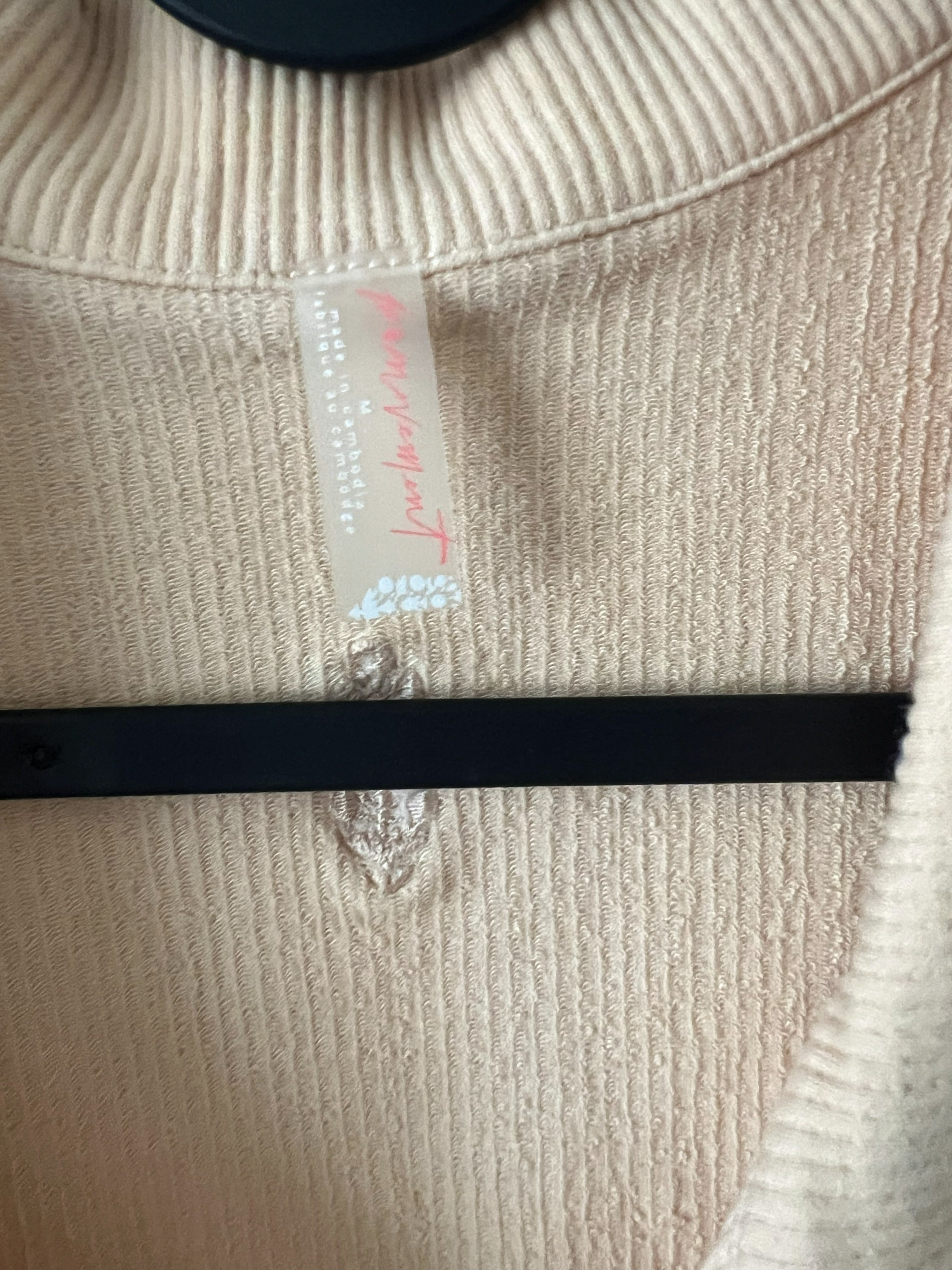 Close-up of a cream-colored ribbed knit sweater with a visible 'FP Movement' brand tag and a black hanger bar across the chest.