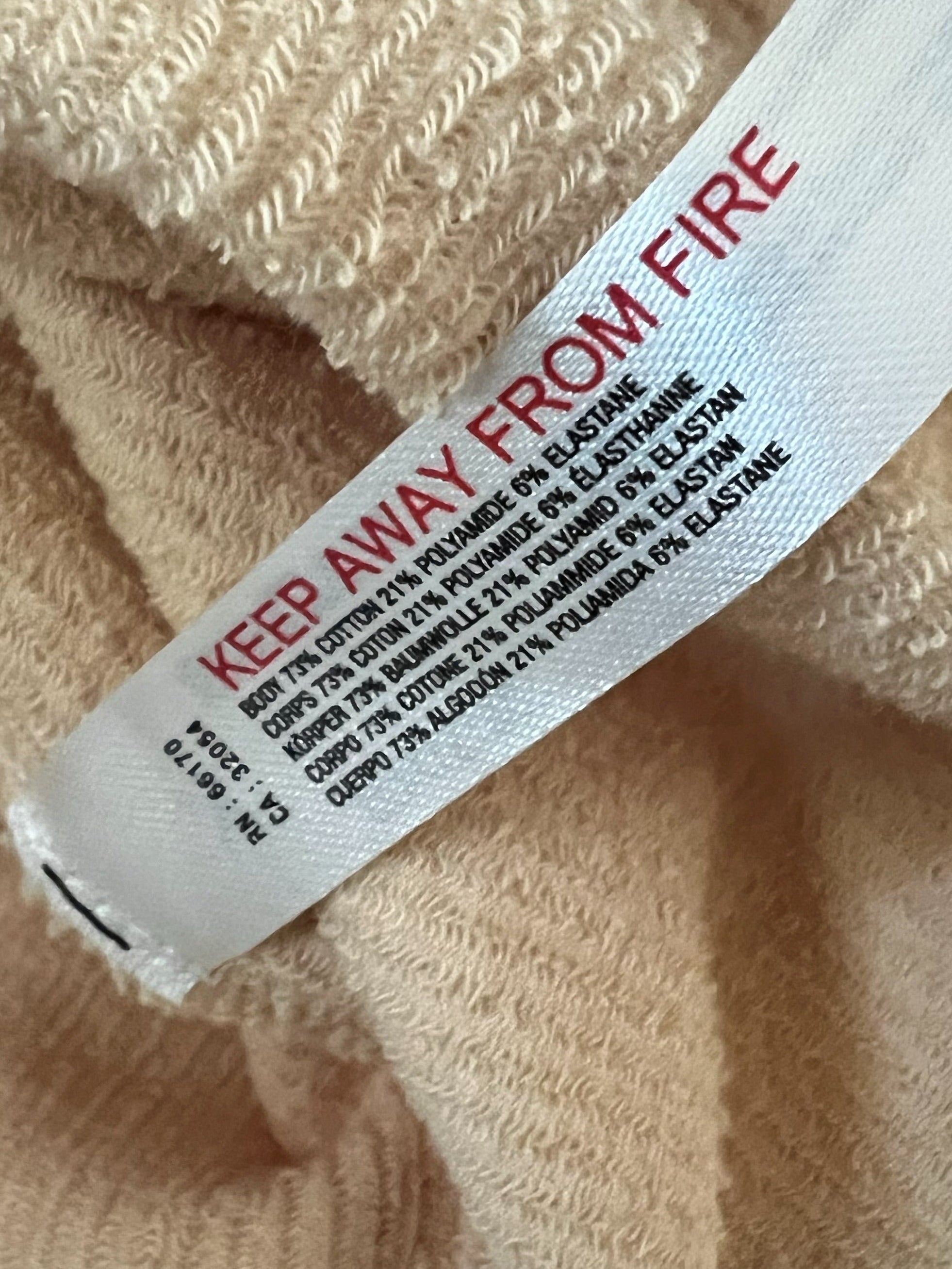 Close-up of a beige ribbed knit garment with a white fabric tag printed with red and black text reading 'KEEP AWAY FROM FIRE'.