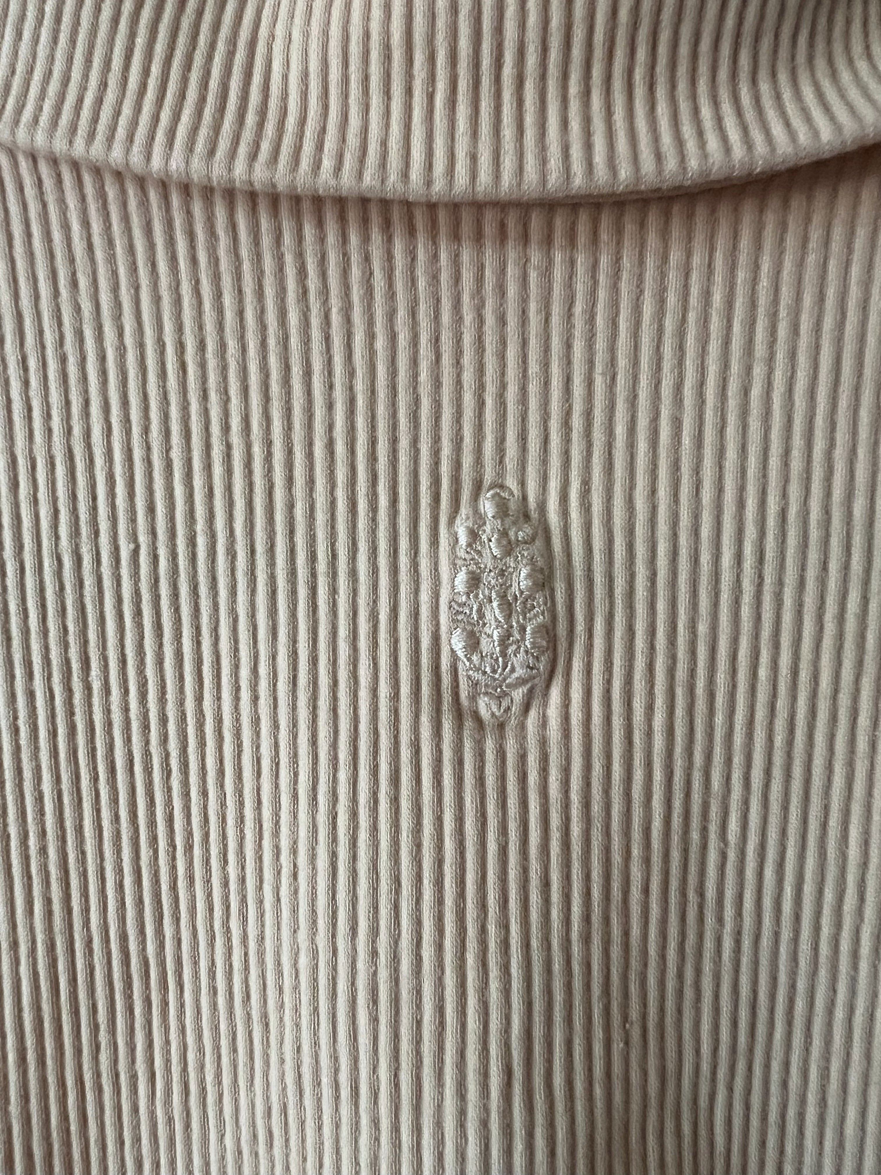 Close-up of a ribbed knit turtleneck sweater in a light beige or cream color, featuring a small embroidered floral crest logo on the chest.