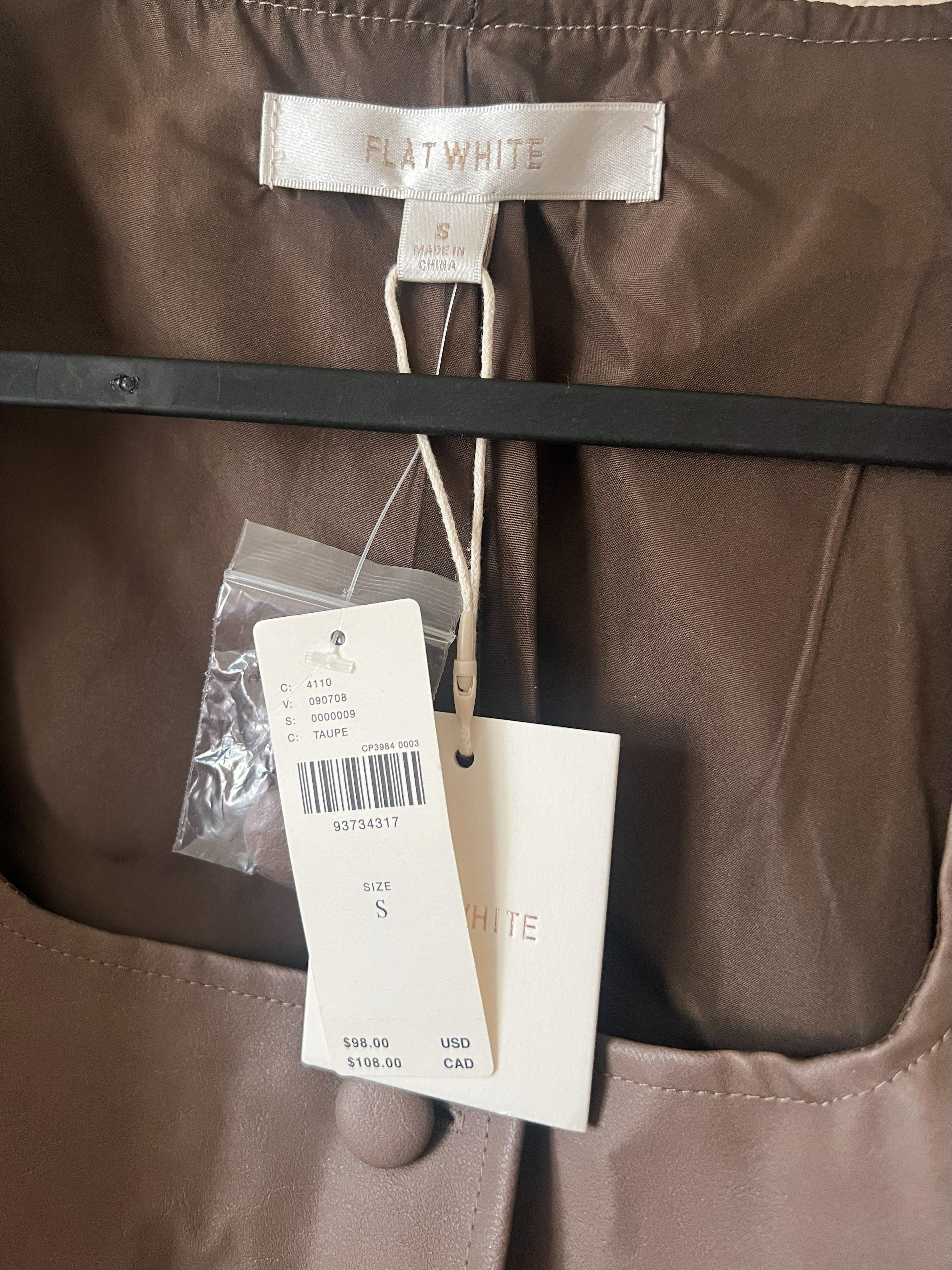 Close-up of a Flat White garment hanging on a black hanger, showing the interior neck label, product tag, and size tag against a brown fabric background.
