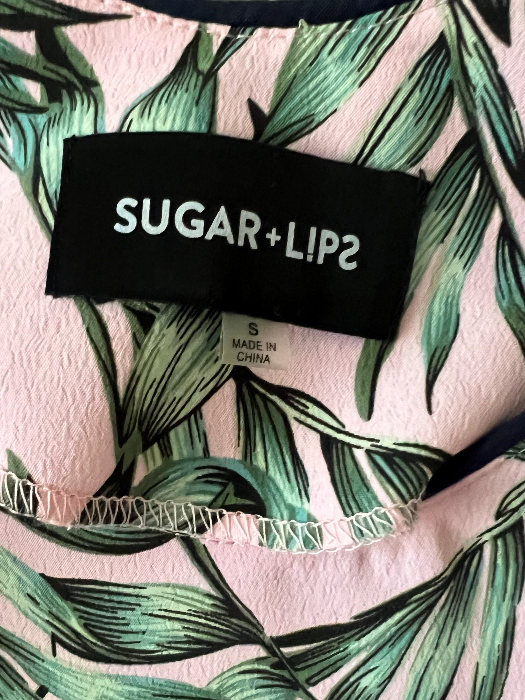 Close-up of a pink garment with a green tropical leaf print and a black brand label reading SUGAR+LIP2.