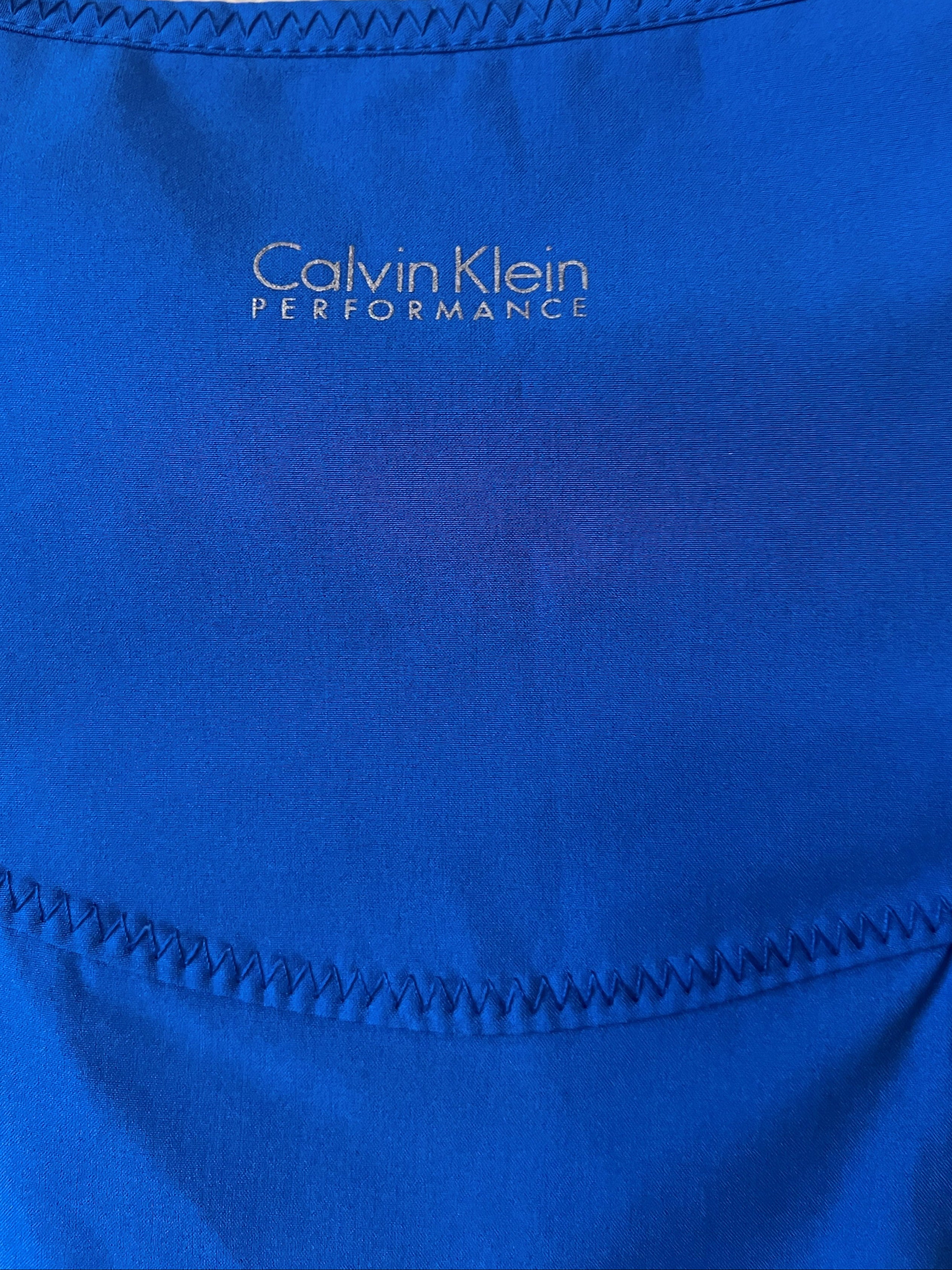 Close-up of a bright blue Calvin Klein Performance athletic top, showing the brand logo and stitched neckline.