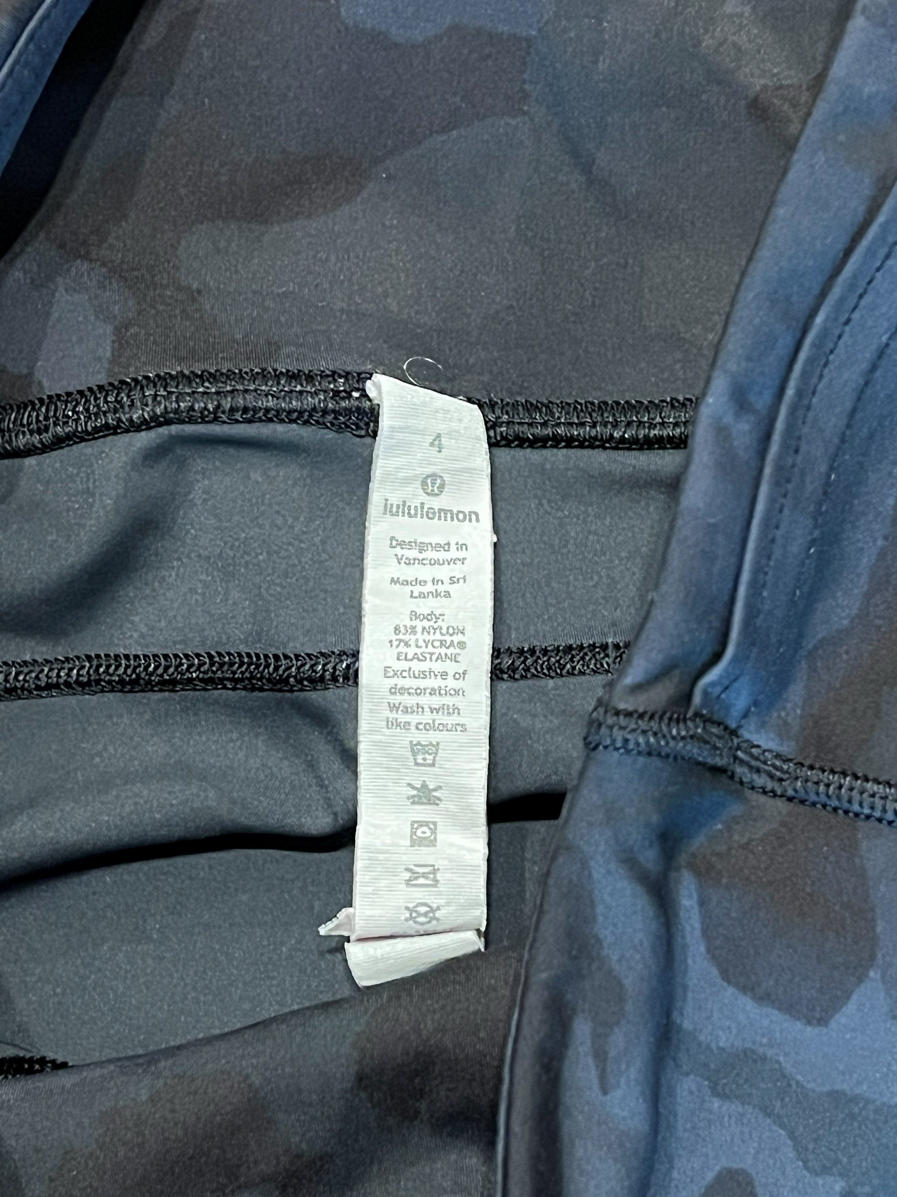 Close-up of a garment's interior neck area showing a white care label with the brand name 'Lululemon' printed on it, alongside black drawstrings and a dark gray camouflage pattern.