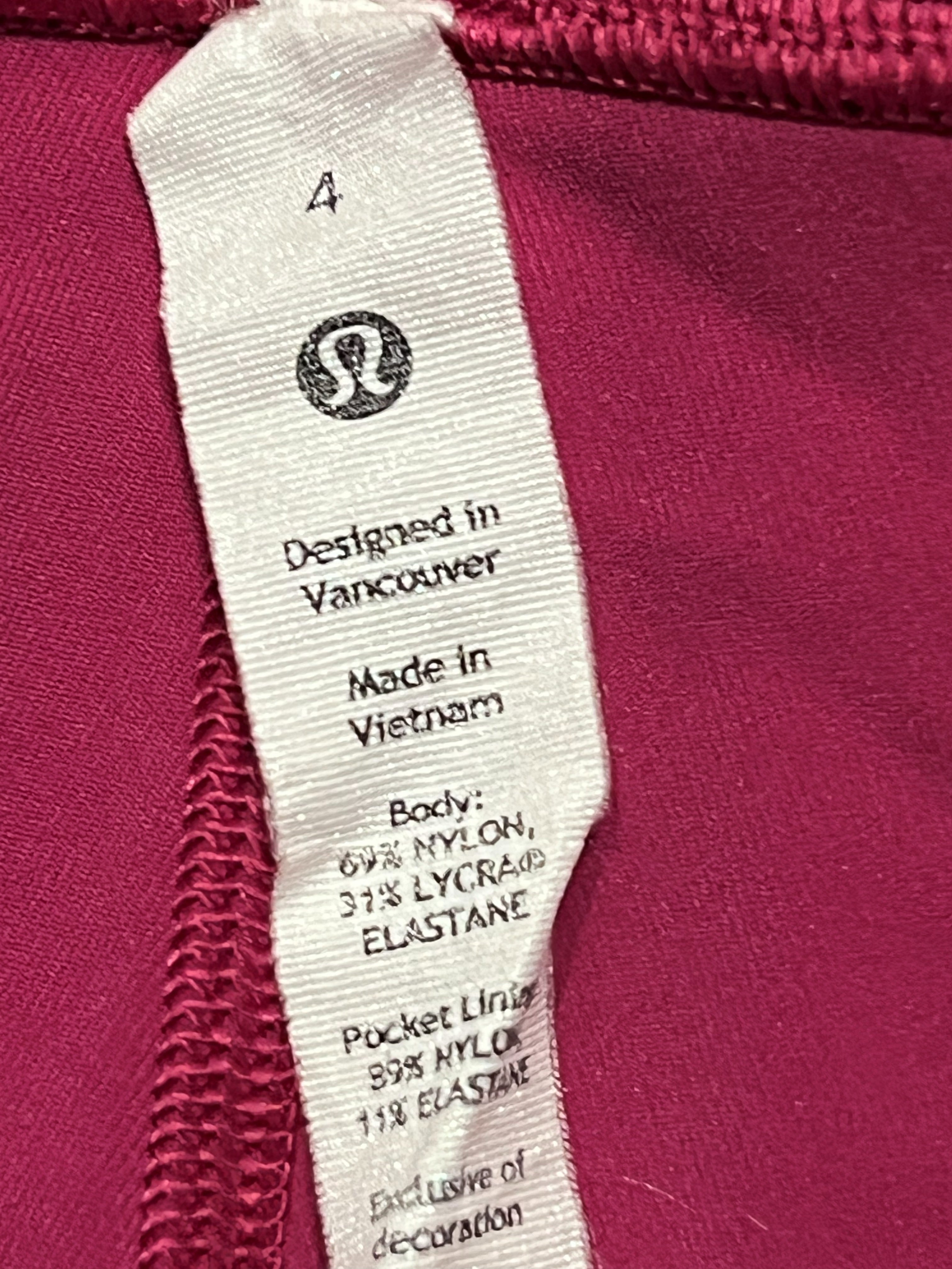 Close-up of a garment's interior neck label showing size 4, 'Designed in Vancouver', 'Made in Vietnam', and the S-Luxe logo, with a deep magenta fabric background.