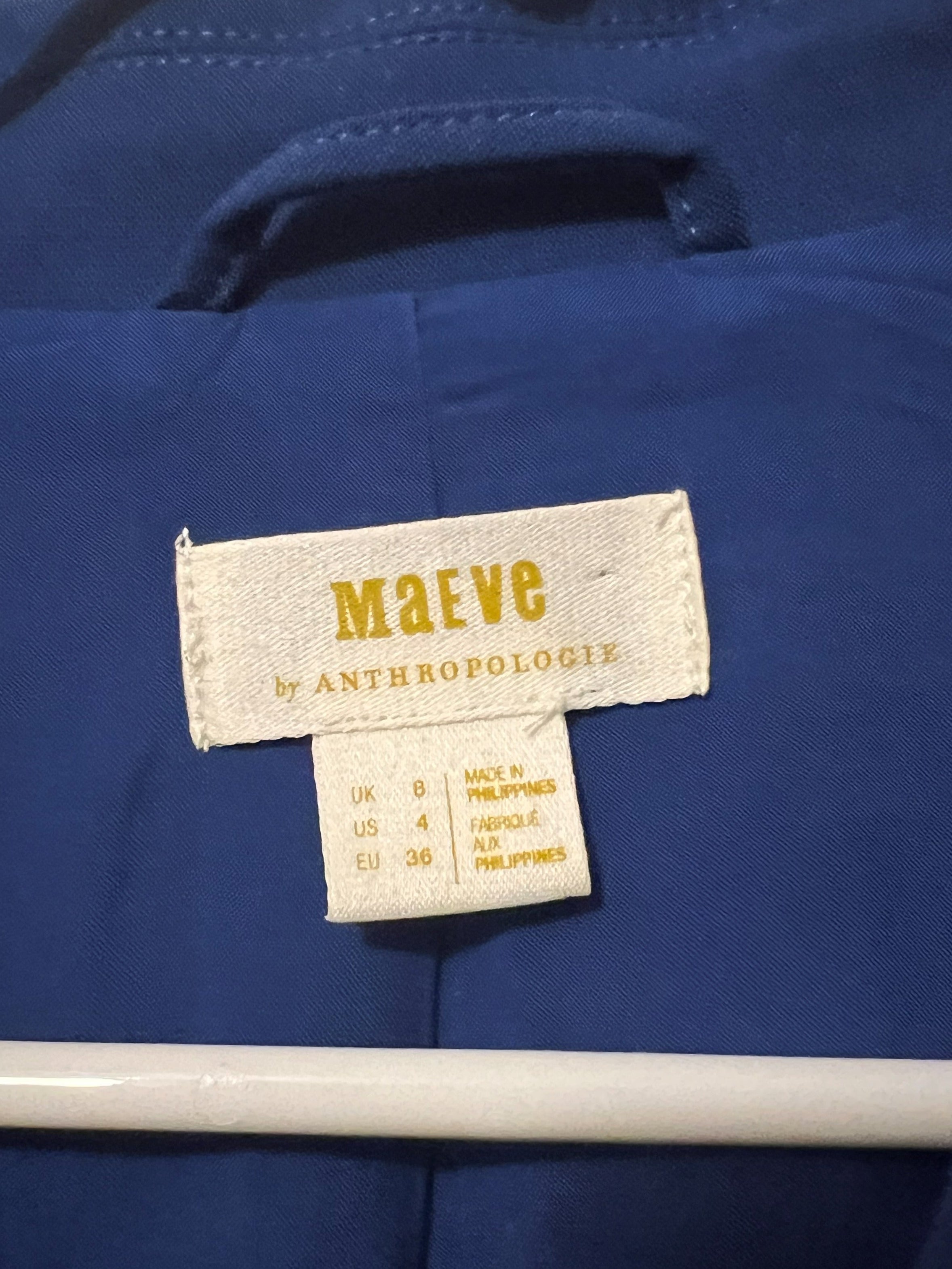 Close-up of a blue garment hanging on a white hanger, showing a sewn-in label that reads 'Maeve by Anthropologie'.