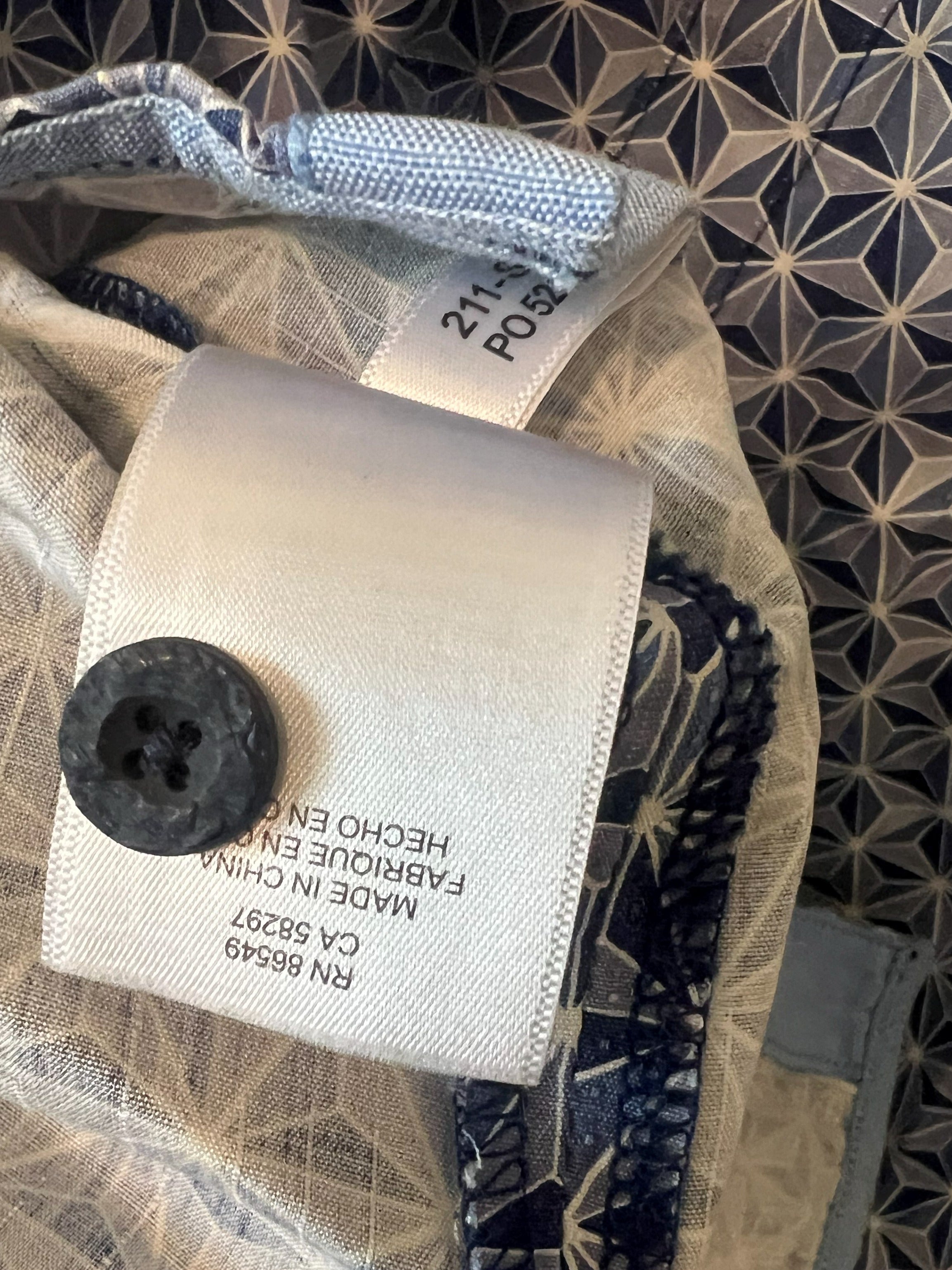 Close-up of a garment's inner neck area showing a white fabric label with a black button, a size tag reading 21 US, and a patterned lining in beige and navy blue.