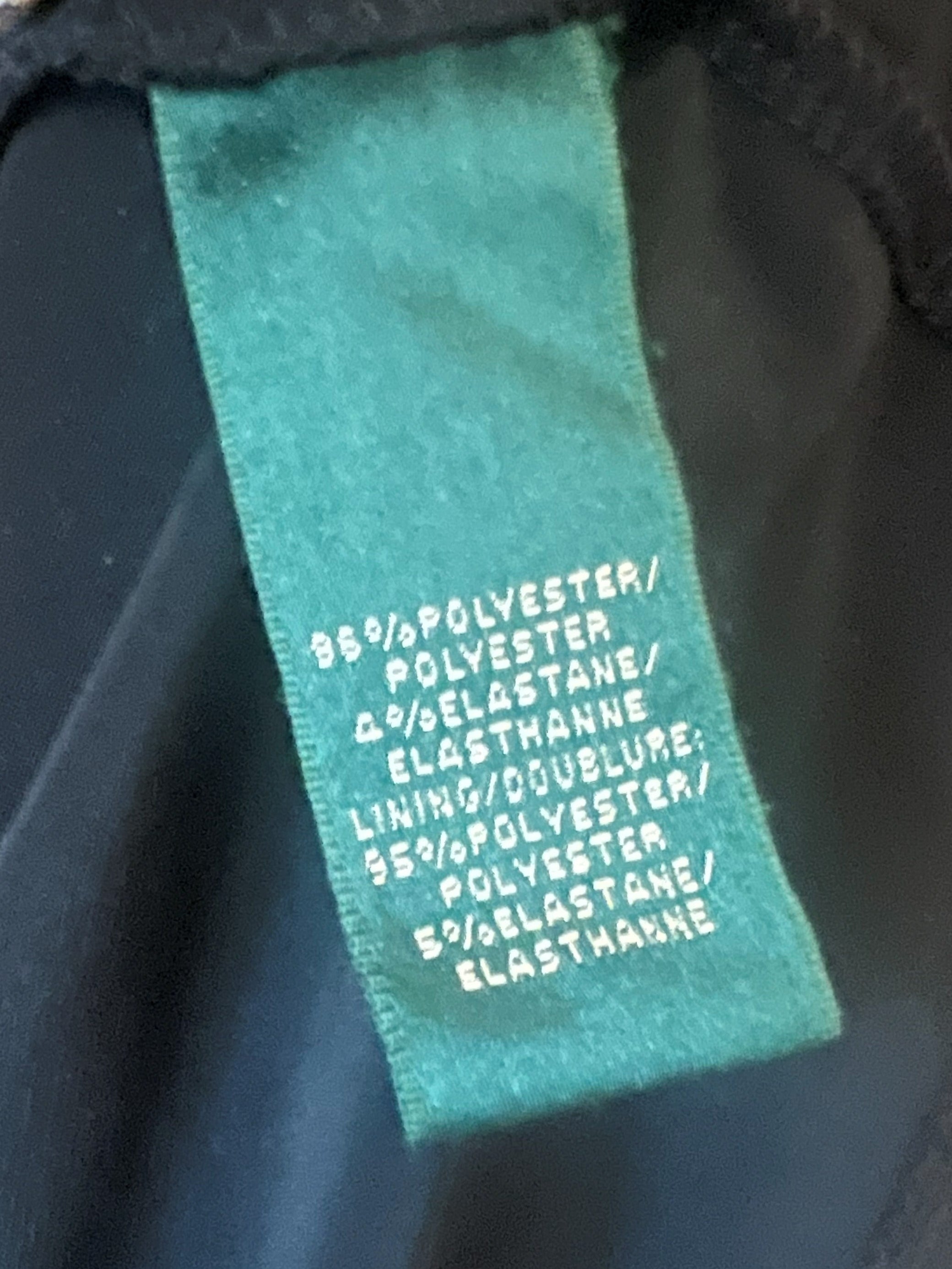 Close-up of a garment's interior neck label made of teal-green fabric with white text reading '95% POLYESTER / POLYESTER / LINING / POLYESTER / ELASTHANNE'.