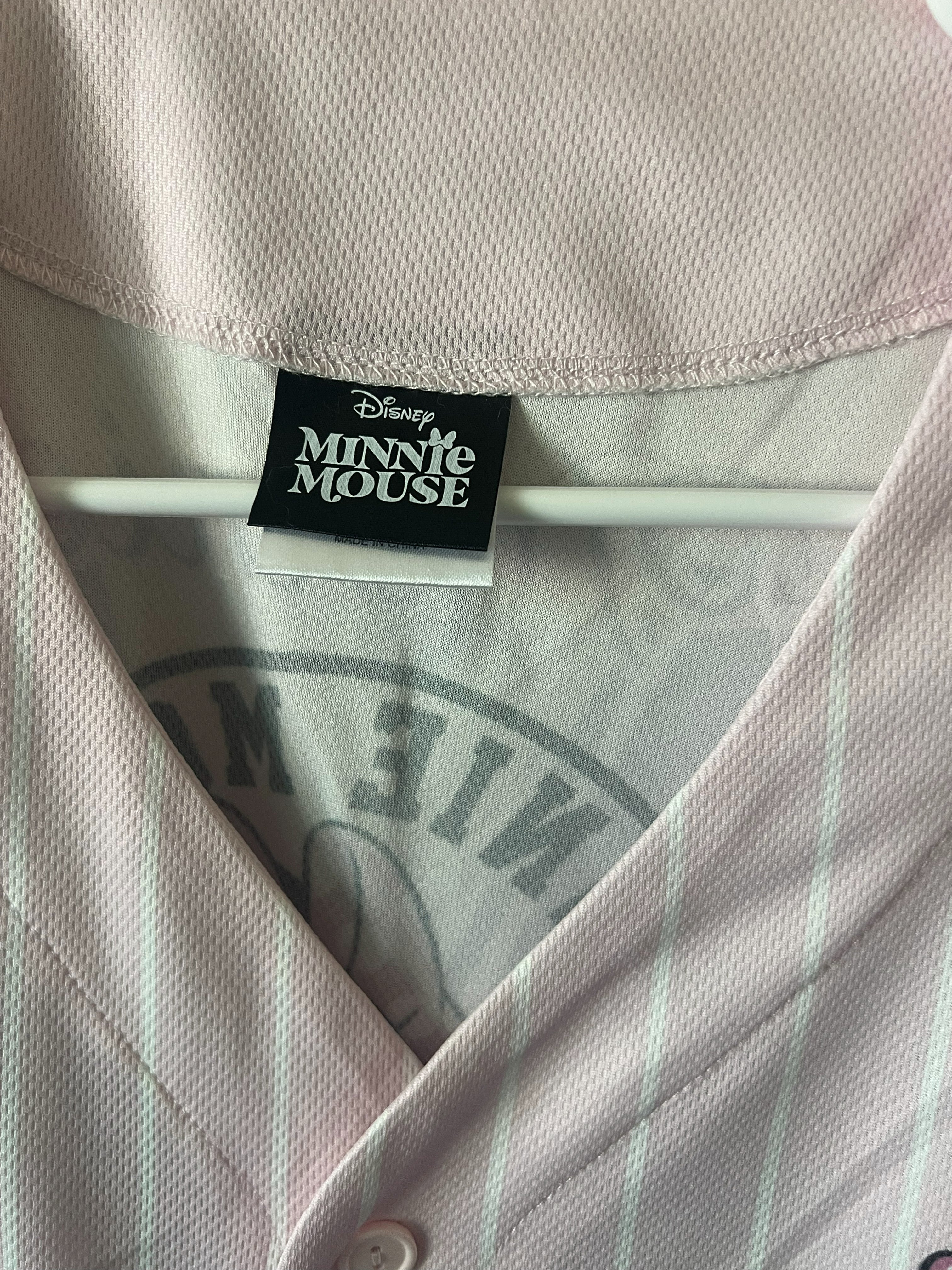 Close-up of a Disney Minnie Mouse button-front shirt in a pale pink color with thin white vertical pinstripes, showing the black and white Disney Minnie Mouse neck label.