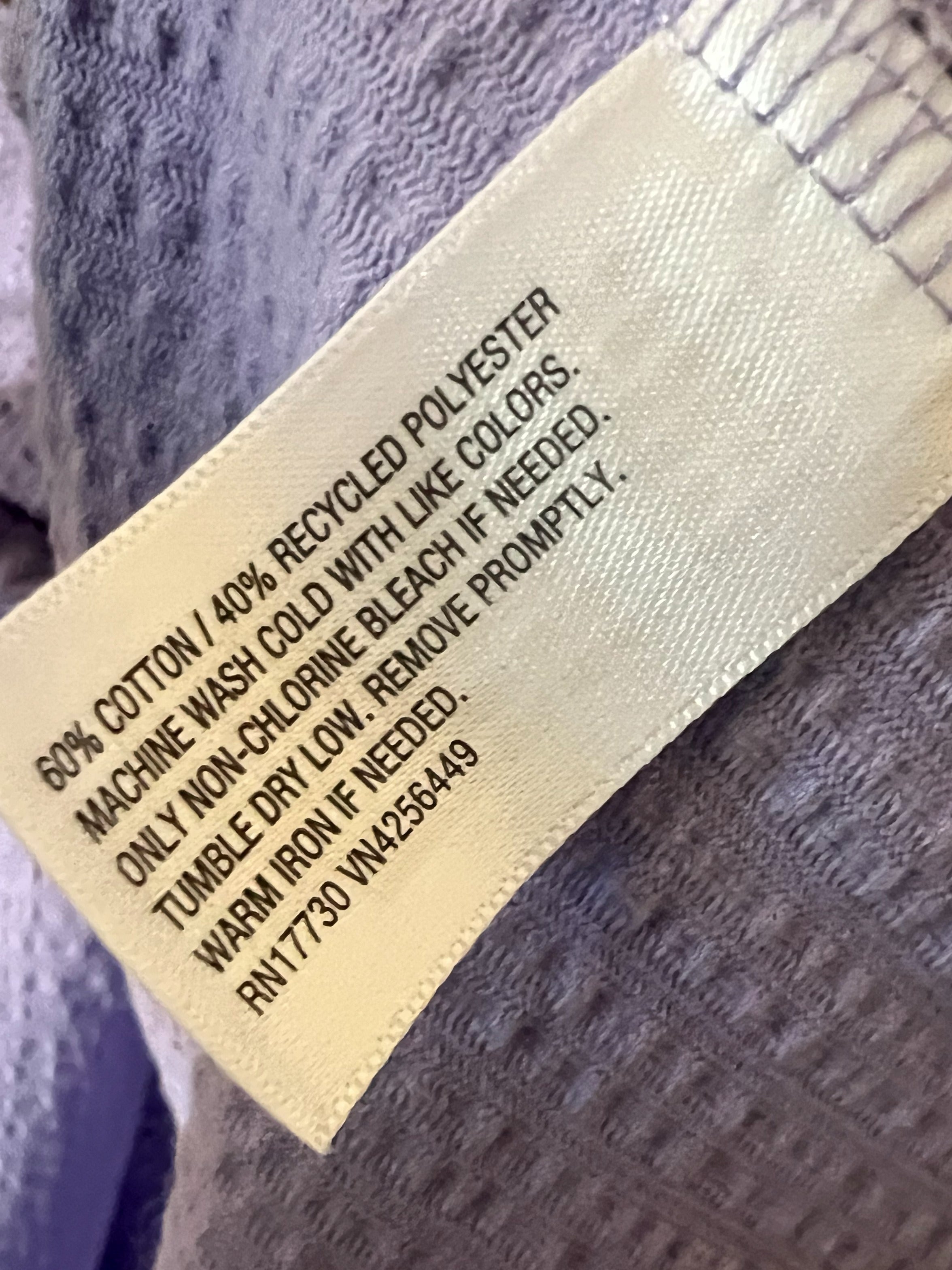 Close-up of a sewn-in product label on a textured lavender-gray fabric, showing text that reads: '60% COTTON / 40% RECYCLED POLYESTER MACHINE WASH COLD WITH LIKE COLORS. ONLY NON-Chlorine Bleach IF FEEDED.