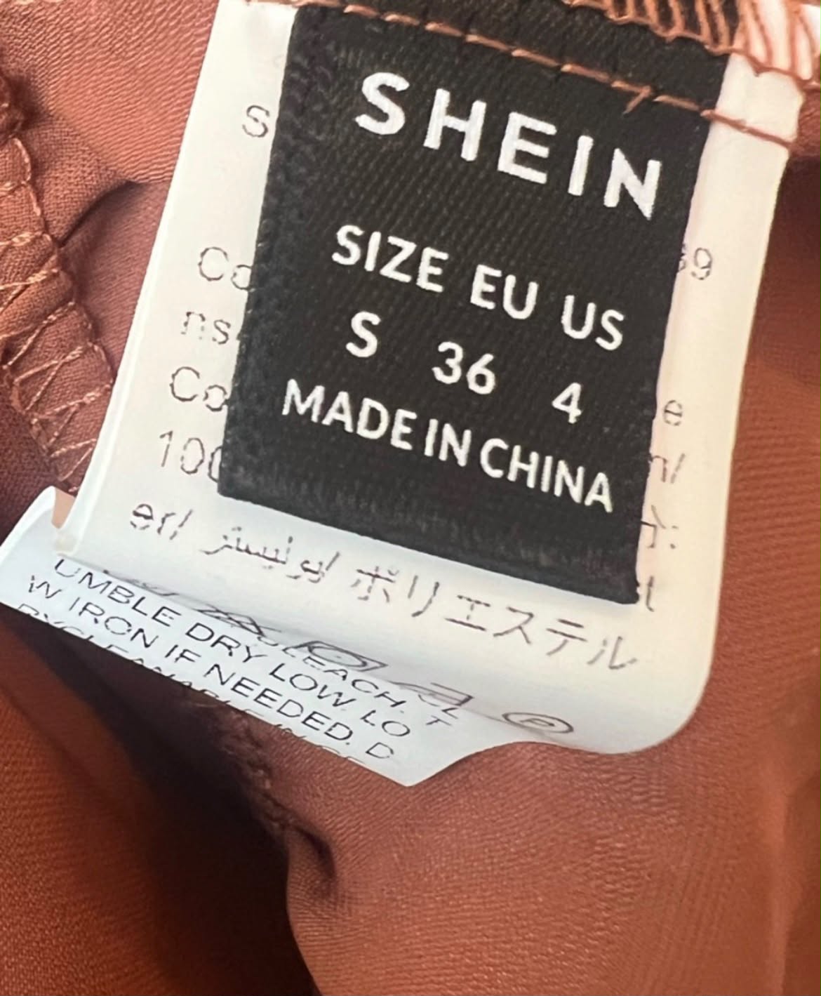 Close-up of a clothing size and brand label reading SHEIN, size EU US S 36 4, Made in China, attached to a rust-brown garment.