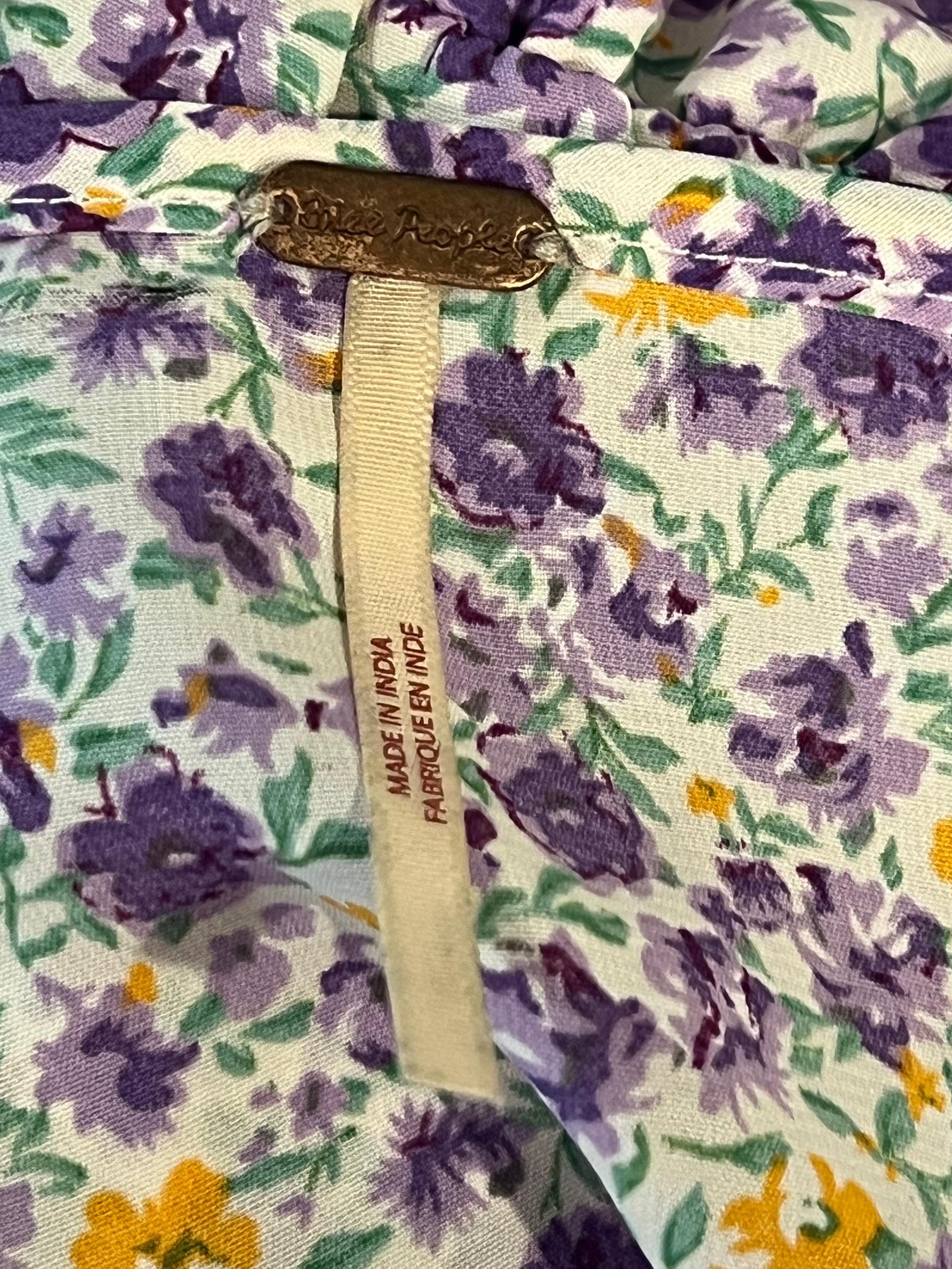 Close-up of a floral fabric garment tag with a purple and yellow floral print on a white background, featuring a beige ribbon label and a brass-colored metal brand plate.