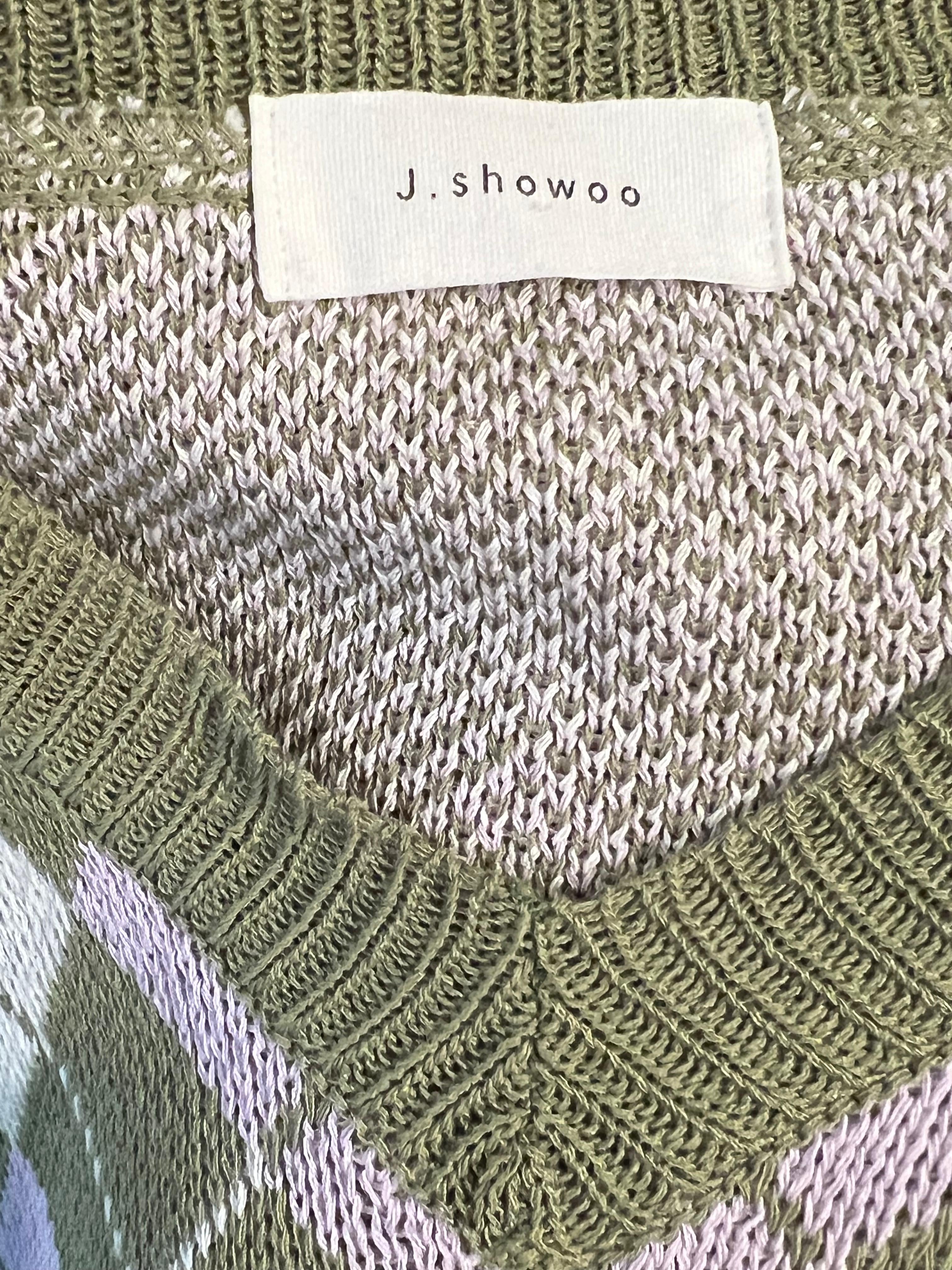 Close-up of a knit sweater showing a ribbed olive green crew neck, a white woven brand label reading J.showoo, and a soft beige-and-white patterned knit body.